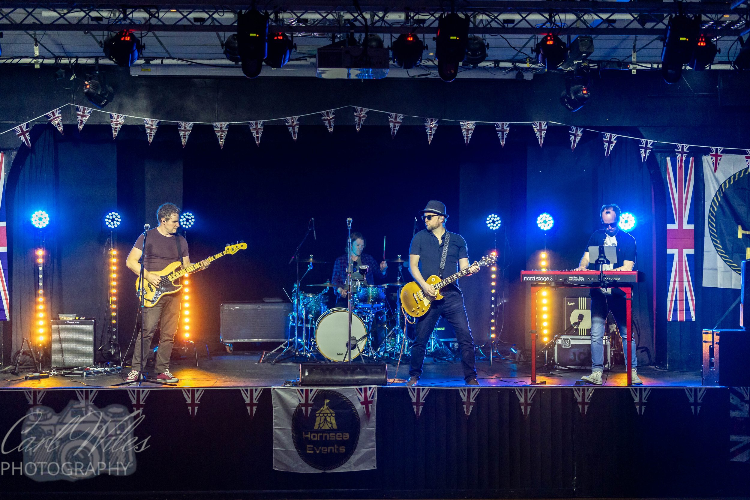 A band performing on stage with a Union Jack banner hanging above. The band includes four members: a bassist, a drummer, a guitarist, and a keyboard player. The stage has colorful lighting and musical equipment, with a Union Jack flag on the right si