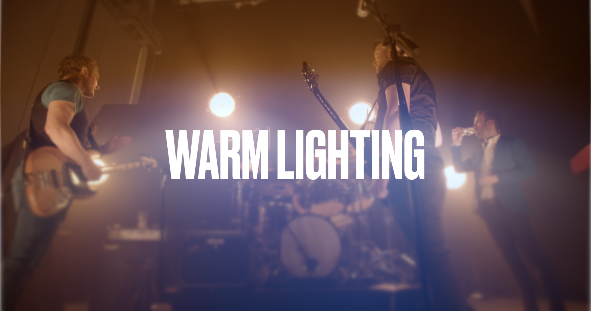 Four musicians performing on stage with warm lighting, one playing guitar, one with a microphone, and two with instruments, under spotlights.