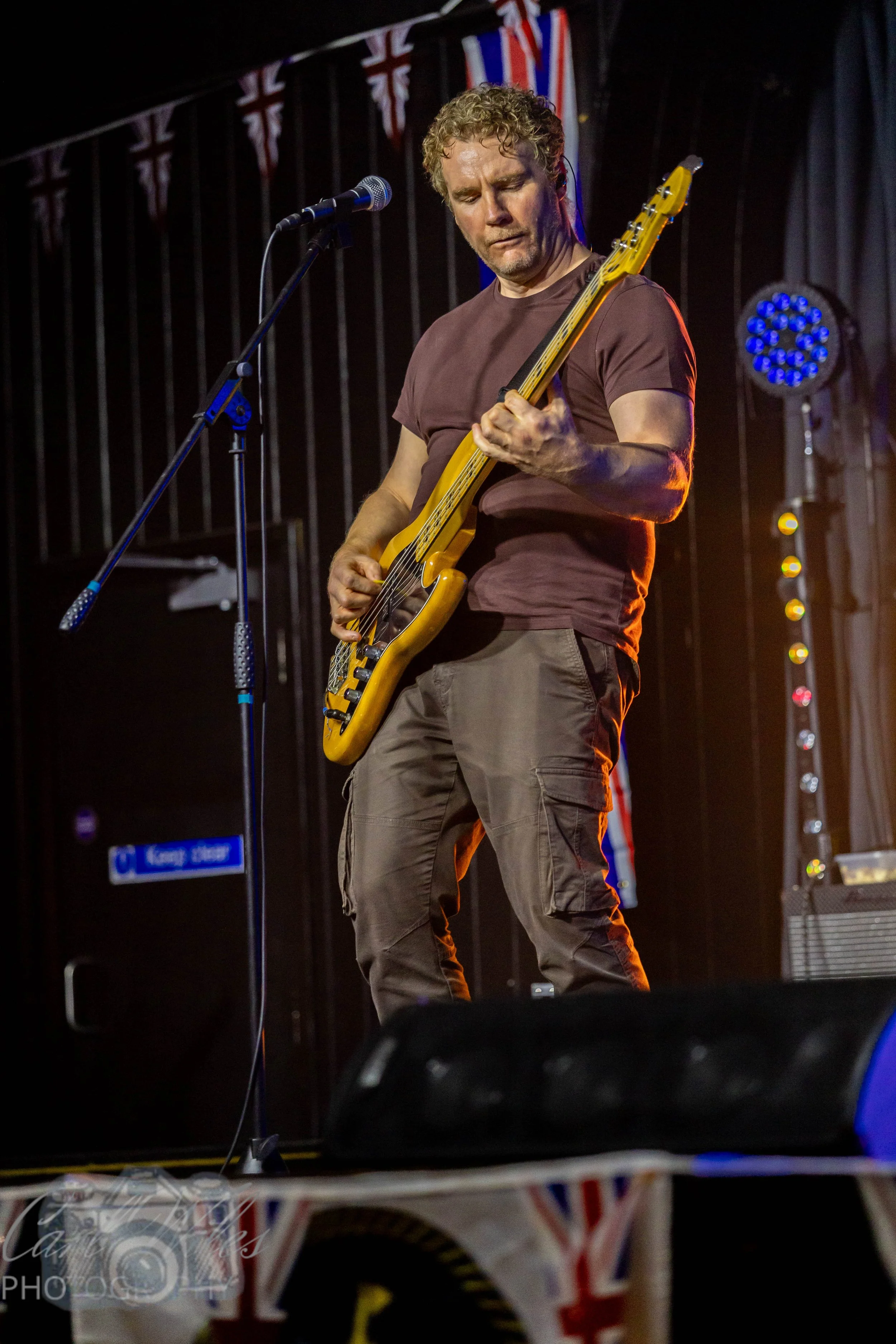A man with curly hair playing an electric bass guitar on stage, with Union Jack flags hanging in the background.