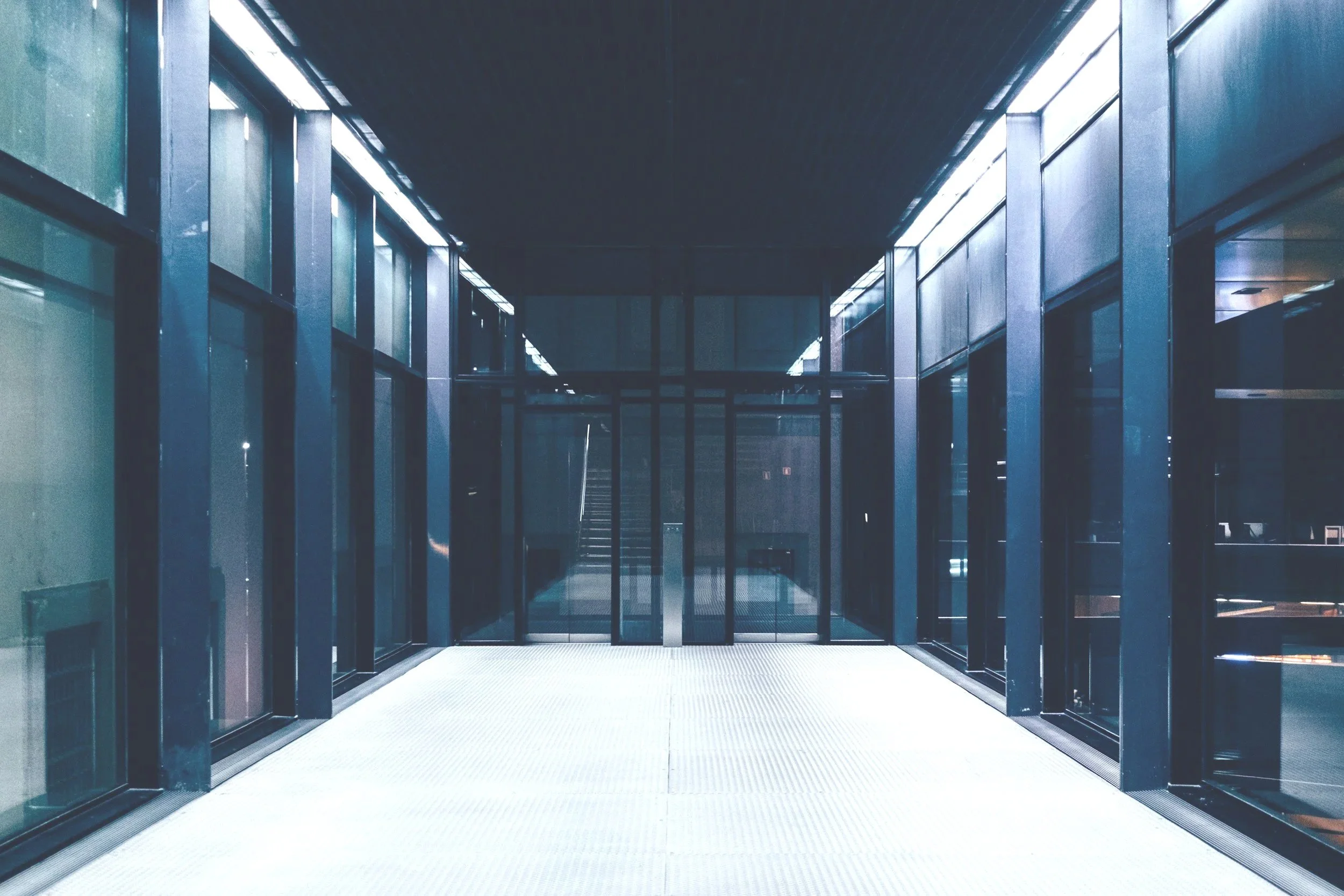 Modern glass corridor with reflective surfaces and a sliding glass door at the end.