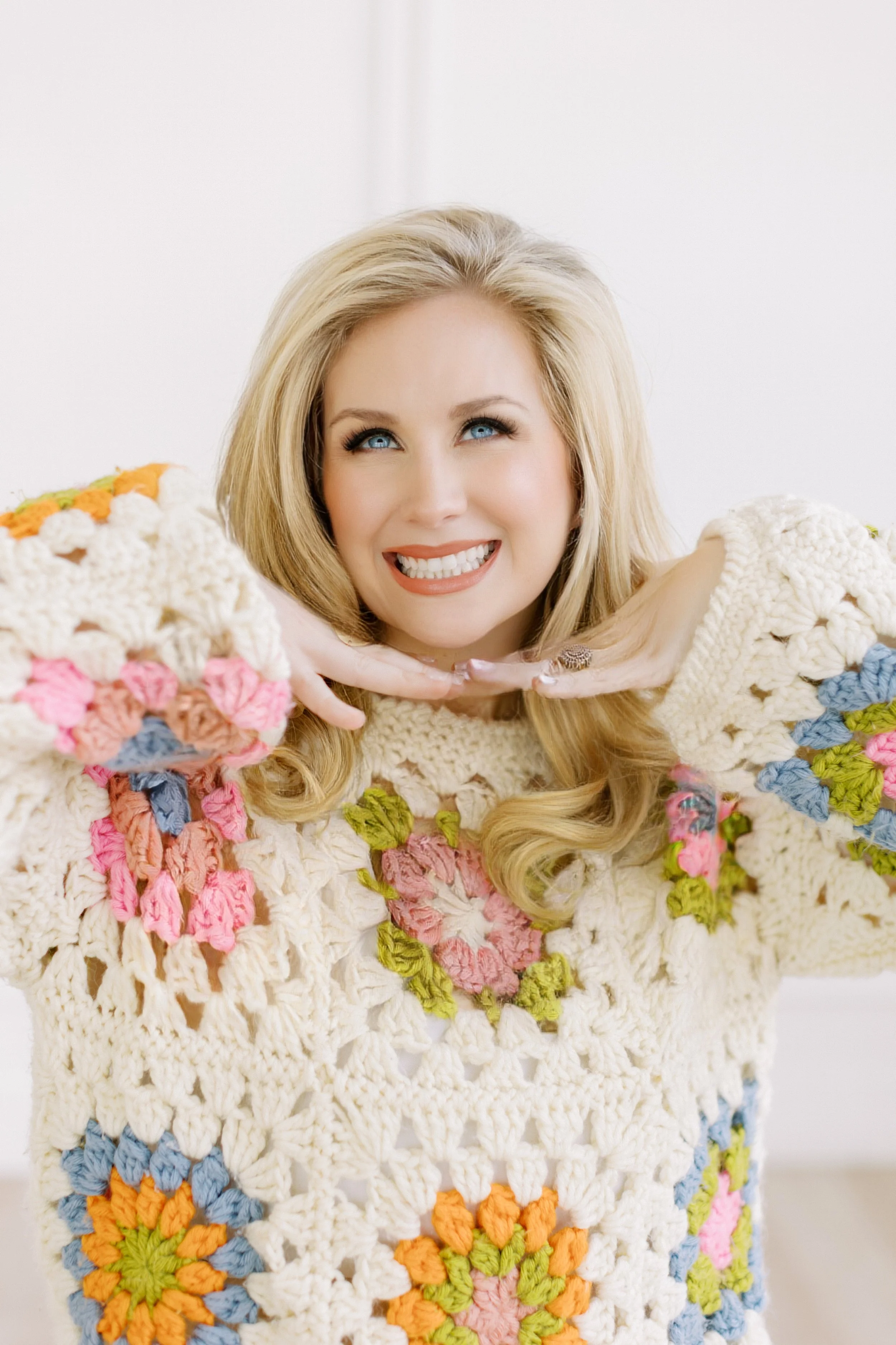 A woman with blonde hair smiling and holding her hands under her chin, wearing a colorful crocheted sweater with flower patterns.