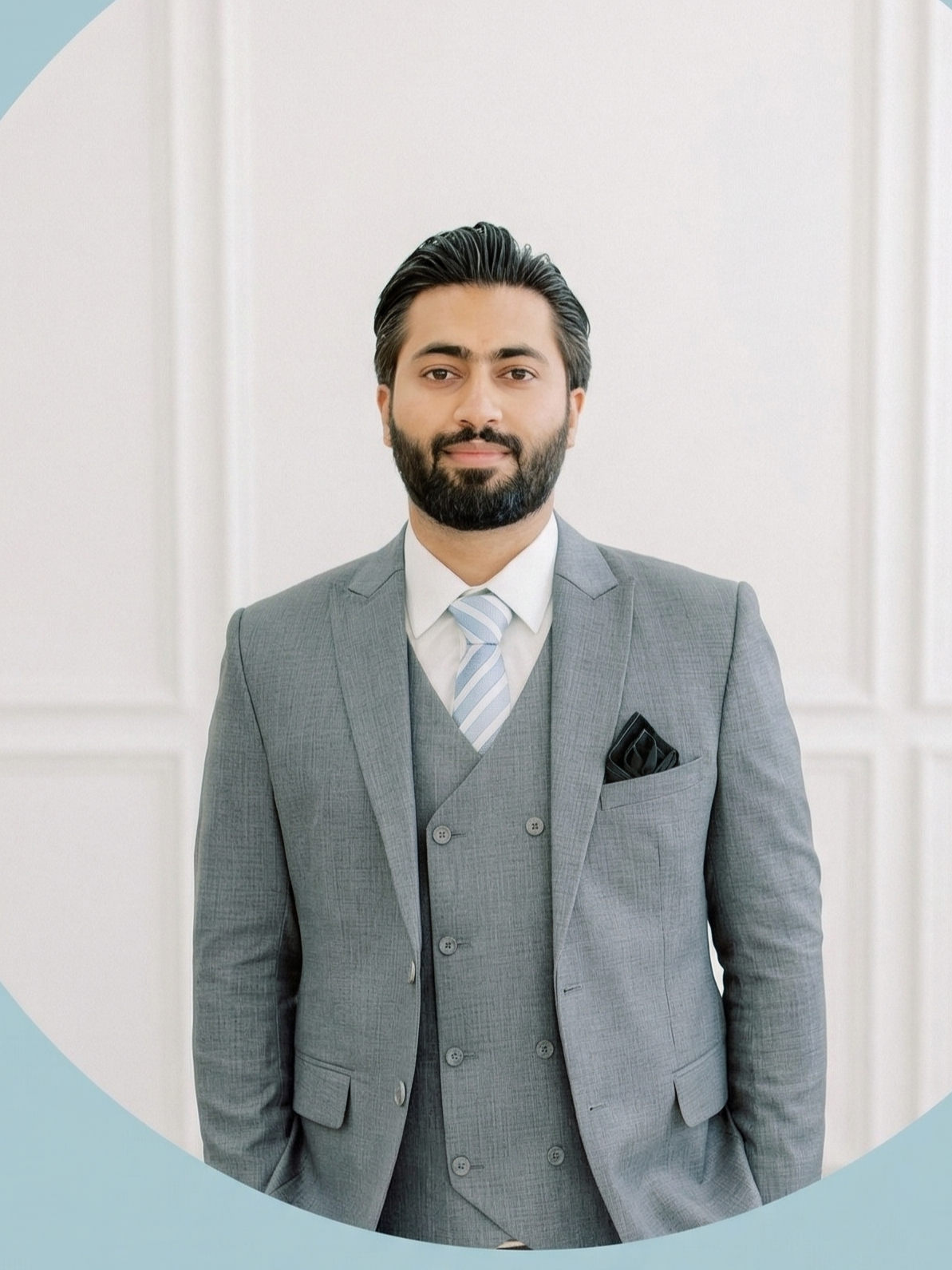 A man with dark hair and a beard wearing a grey suit, white shirt, and striped tie, standing in front of a plain light-colored wall.