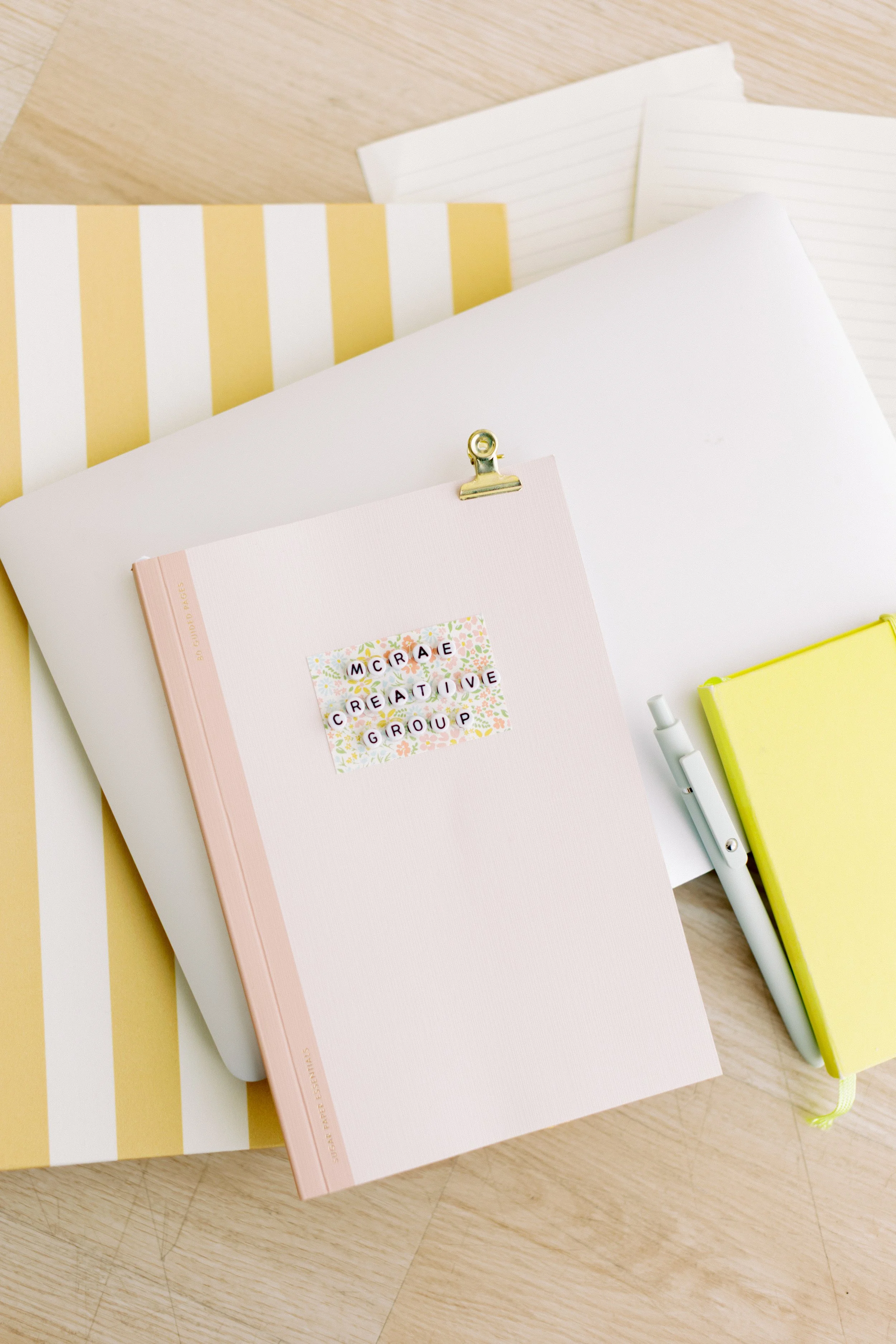 A pink notebook with colorful lettering spelling 'McCrae Creative Group' on the cover, a yellow notebook, a white pen, and sheets of lined paper arranged on a wooden surface.
