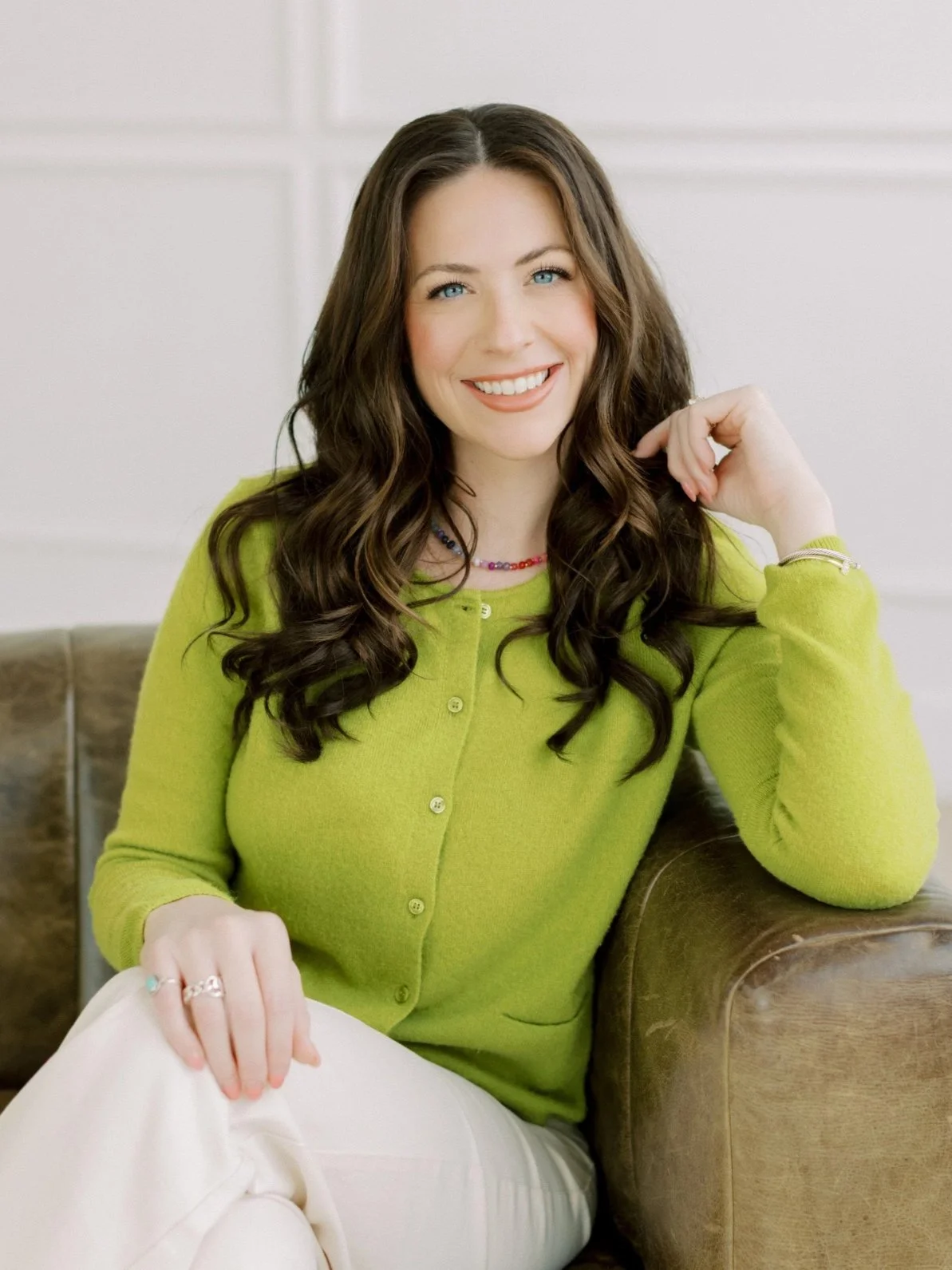 A woman with long, curly brown hair and blue eyes sitting on a brown leather couch, smiling, wearing a bright green cardigan and light-colored pants, in a well-lit room.