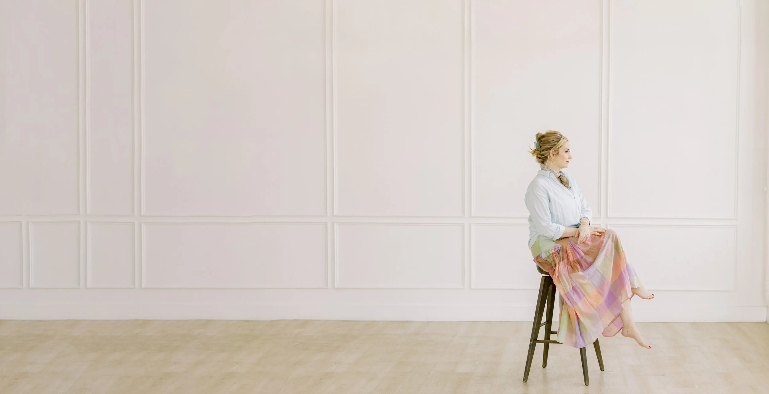 A woman with blonde hair in a bun, wearing a light blue shirt and a pastel-colored skirt, sitting sideways on a wooden stool in a bright, minimalist room with white walls and light wood flooring.