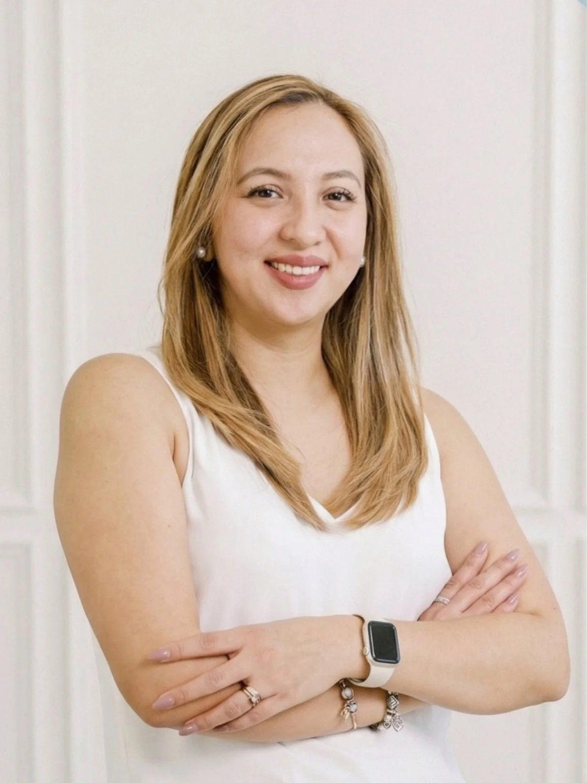 A woman with blond hair in a white sleeveless top standing with her arms crossed, smiling against a plain white background.