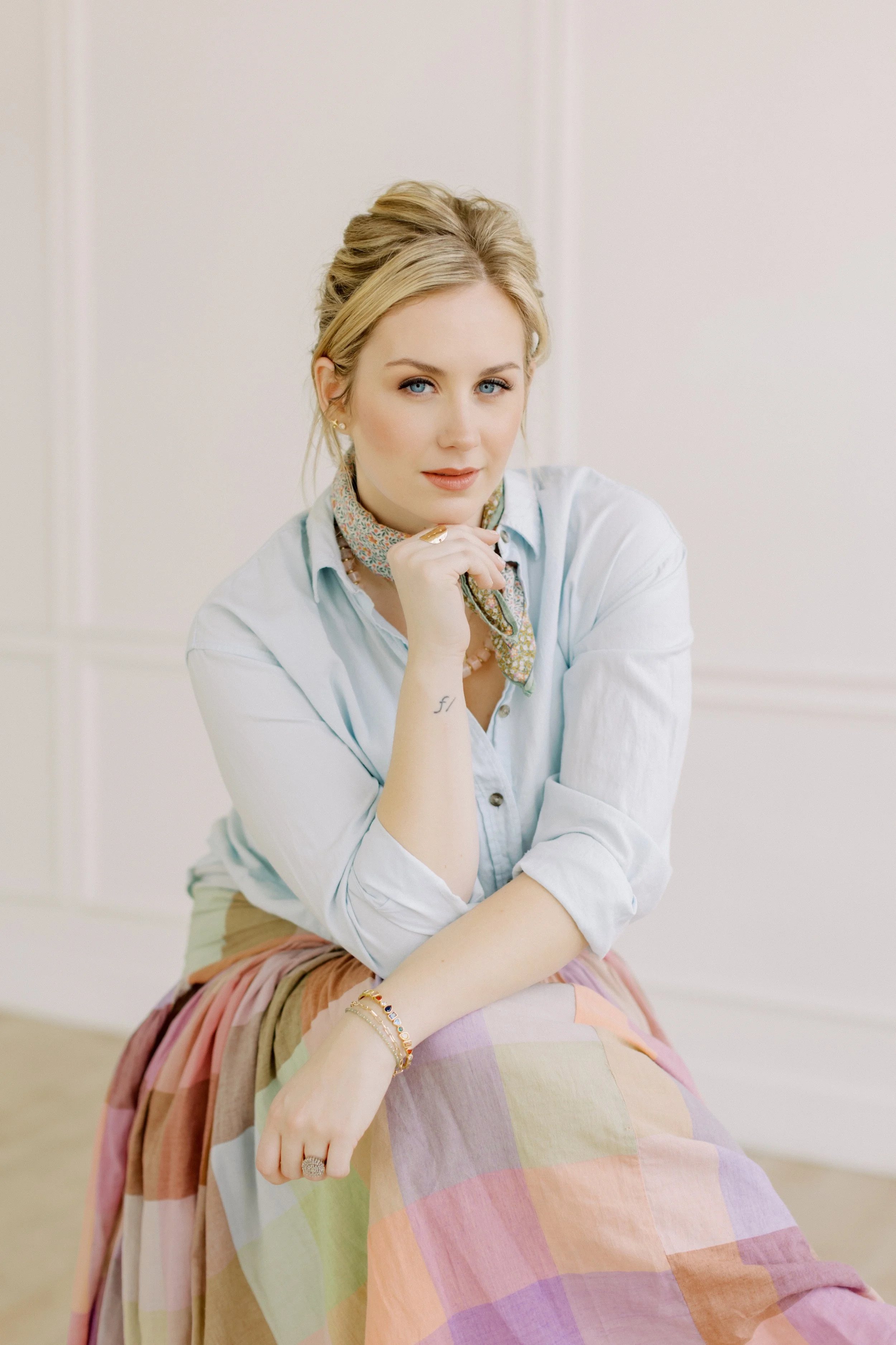 Lauren McRae with blonde hair, blue eyes, and wearing a light blue blouse, a colorful scarf, and a pastel-colored skirt, sitting indoors with her chin resting on her hand and looking at the camera.