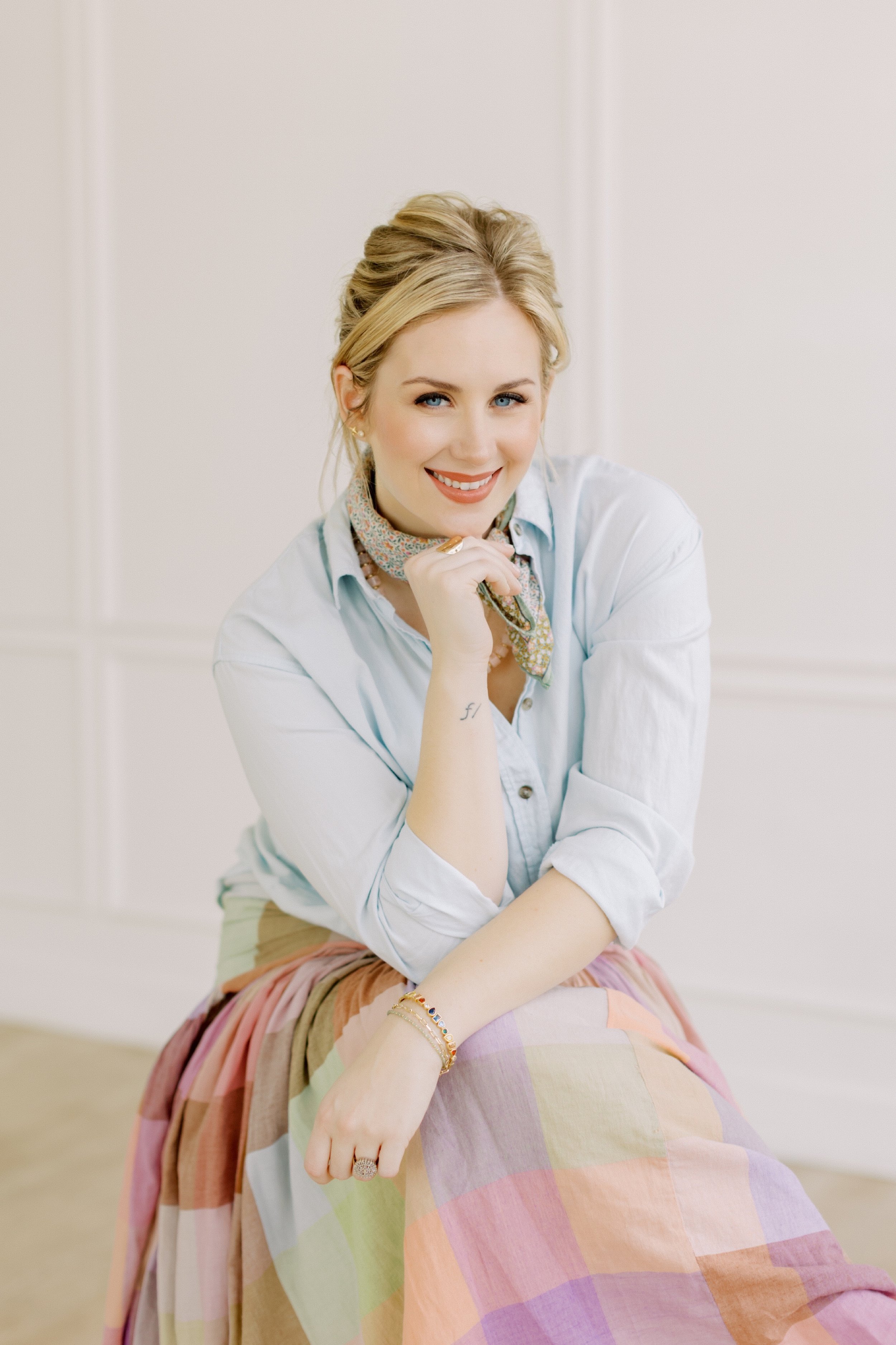 A woman with blonde hair styled in an updo, wearing a light blue button-up shirt, a colorful patterned scarf, and a multicolored pastel skirt, sitting on the floor and smiling at the camera.