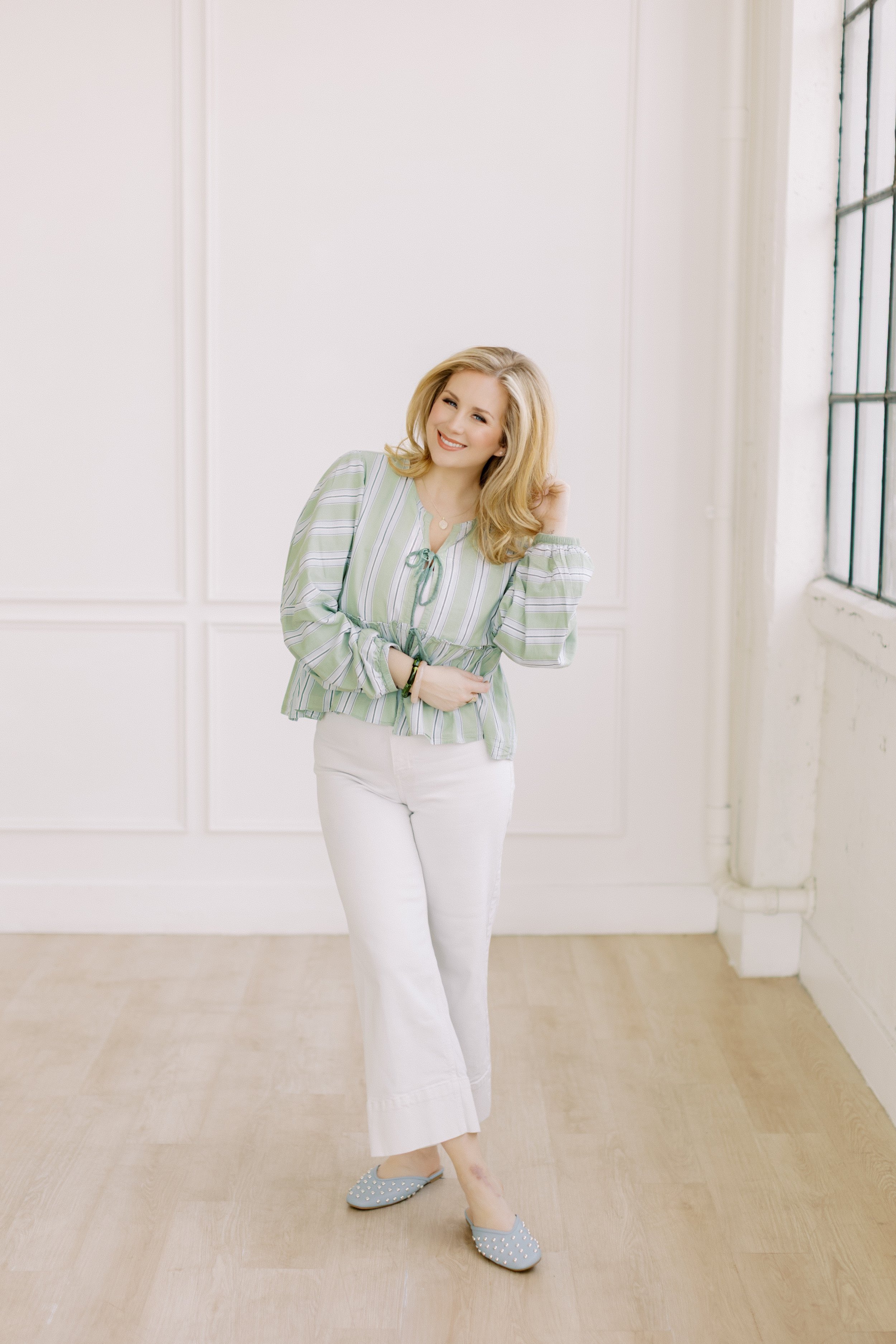Lauren McRae with blonde hair smiling, wearing a green and white striped blouse, white pants, and blue studded shoes, standing in a bright room with white walls and a large window.