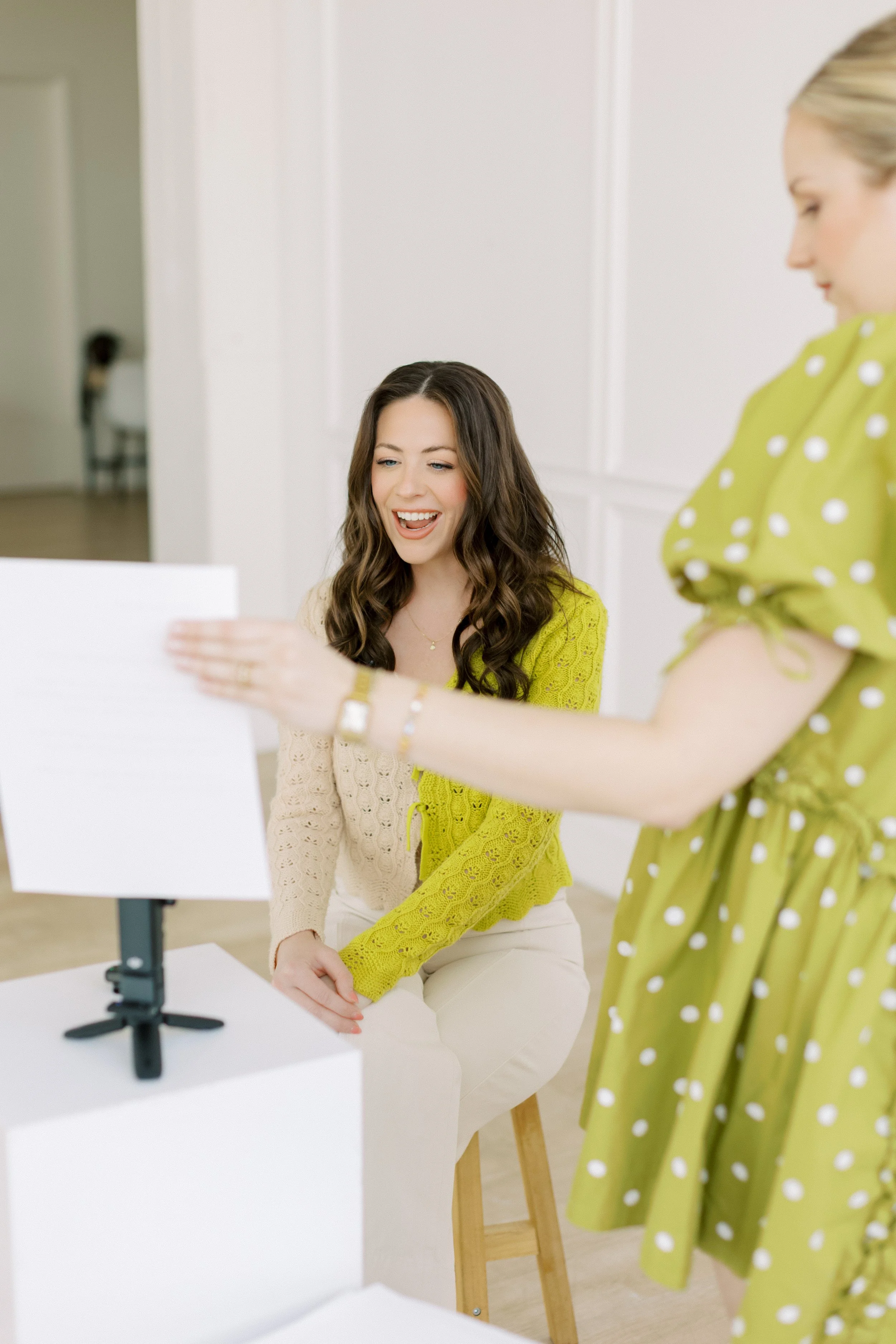 A woman with long brown hair, wearing a cream-colored sweater and cream pants, sitting on a stool, smiling as she looks at a mirror or screen being held by another woman with blonde hair, wearing a green polka-dot dress in a bright, white room.