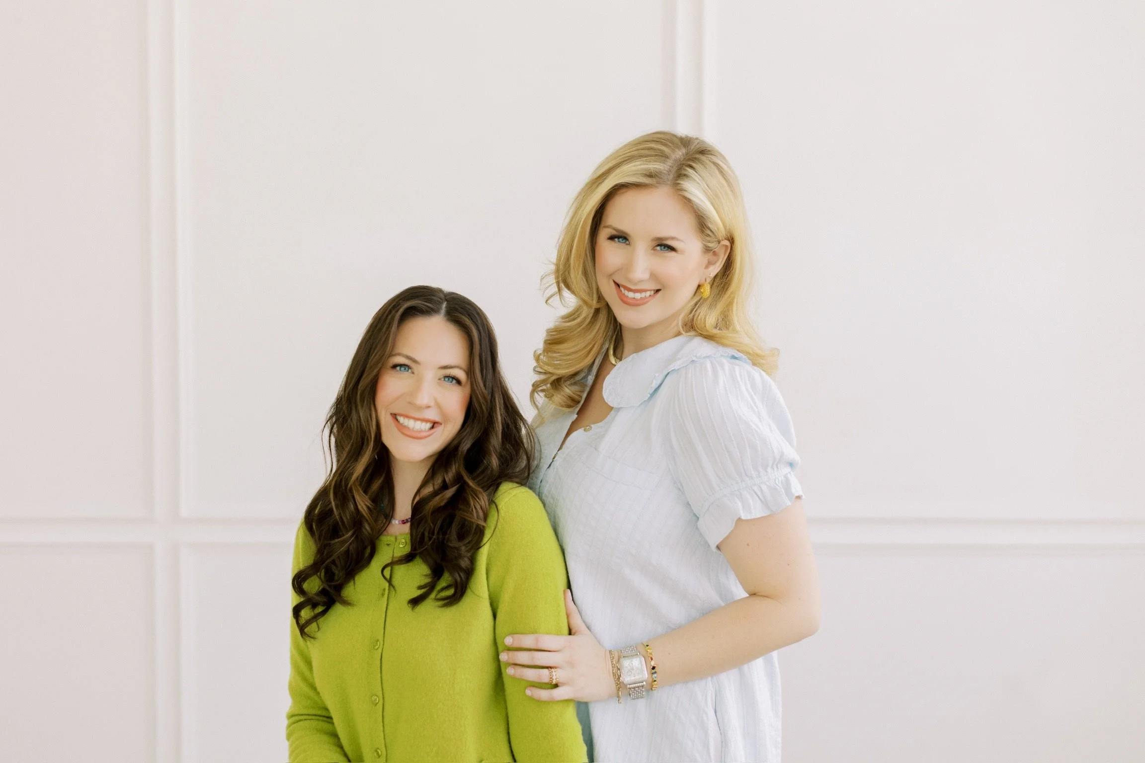 Two women smiling and standing close together, one with long brown hair wearing a green shirt and the other with blonde hair wearing a white shirt, in a bright room with a plain white wall background.