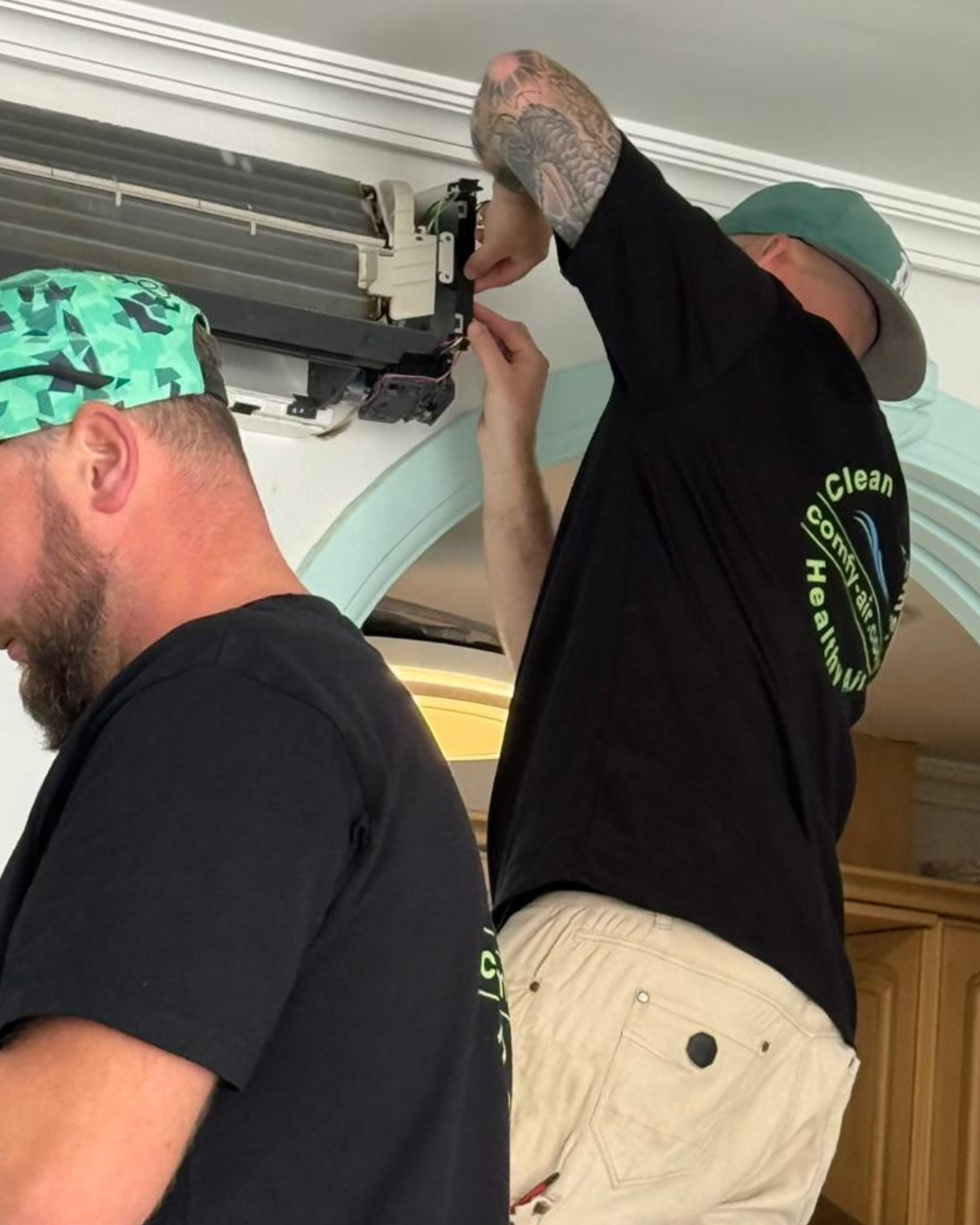 Two men working on an air conditioning unit. One man has a tattooed head and is wearing a black shirt, the other has a tattooed arm and is wearing a hat and beige pants.