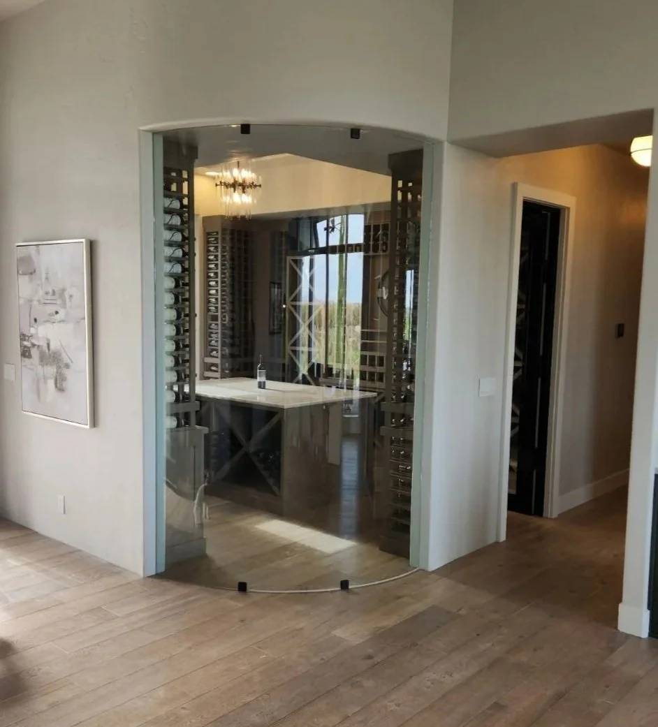 Modern home interior featuring a glass-enclosed wine cellar with wooden wine racks, a small central table, and a view of the outdoors through a window. The room has hardwood flooring and neutral wall colors.
