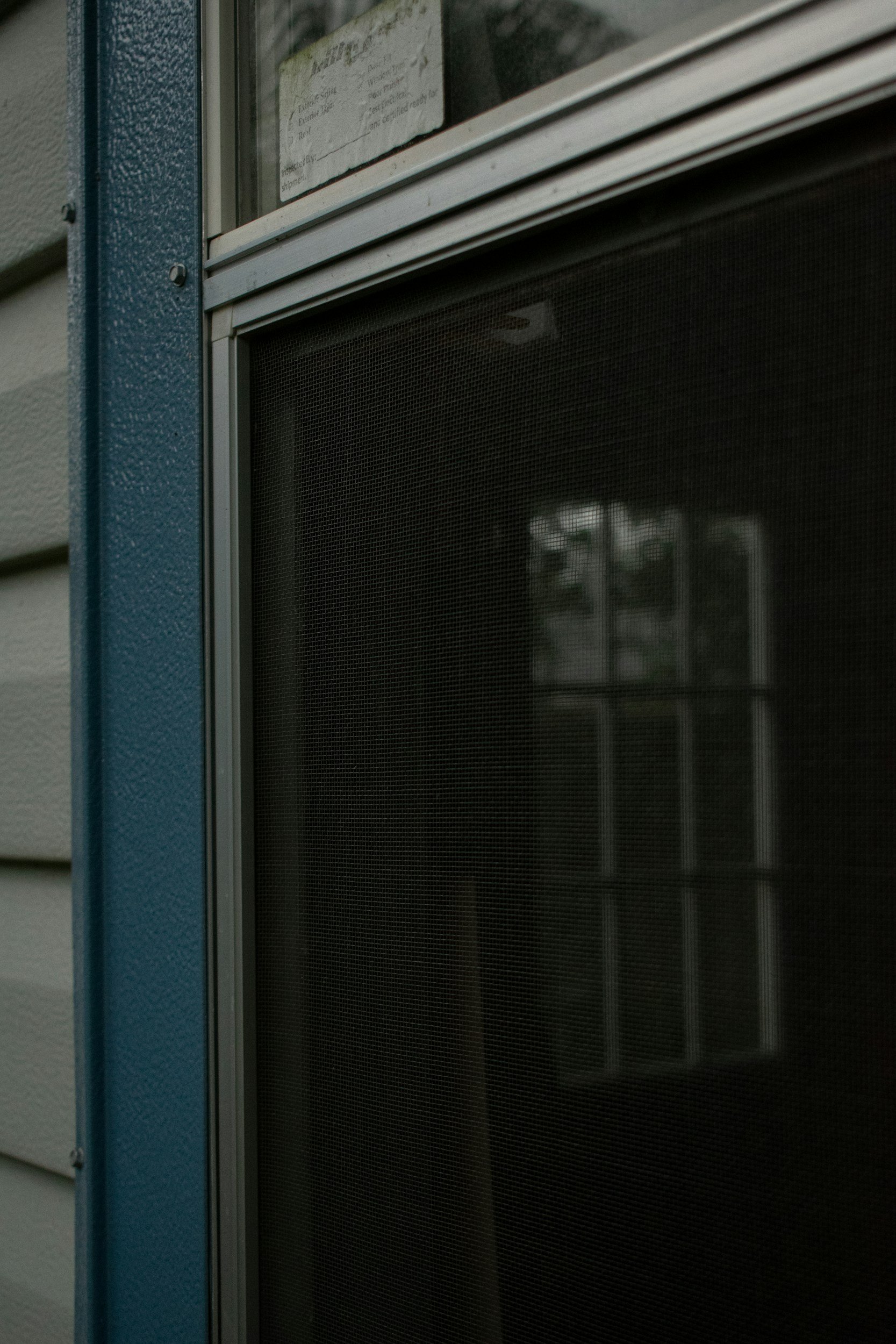 Close-up of a window with a mesh screen, part of a blue and beige house exterior, with reflection of a porch and railing.