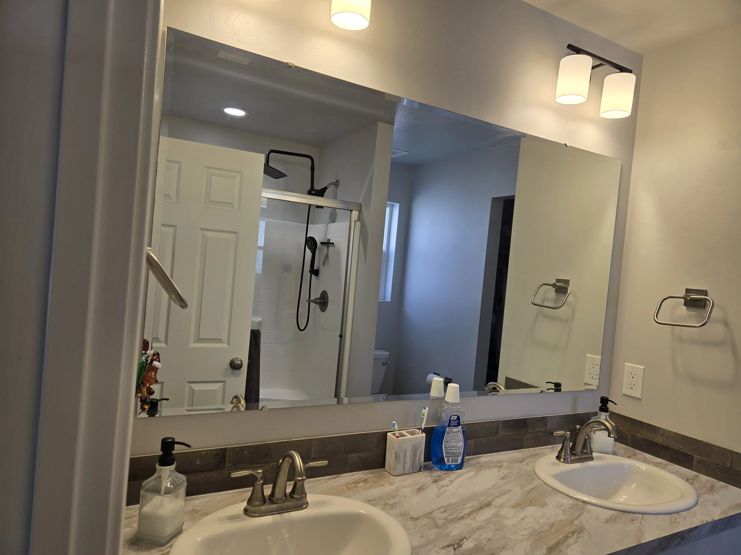 A bathroom countertop with two sinks, soap dispensers, a toothbrush holder, and a mirror reflecting a shower area with a glass door and shower fixtures.