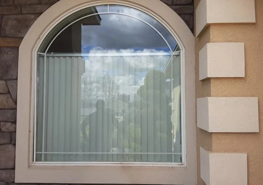 A large arched window with vertical blinds, reflecting clouds, trees, and a person taking a photo outside.