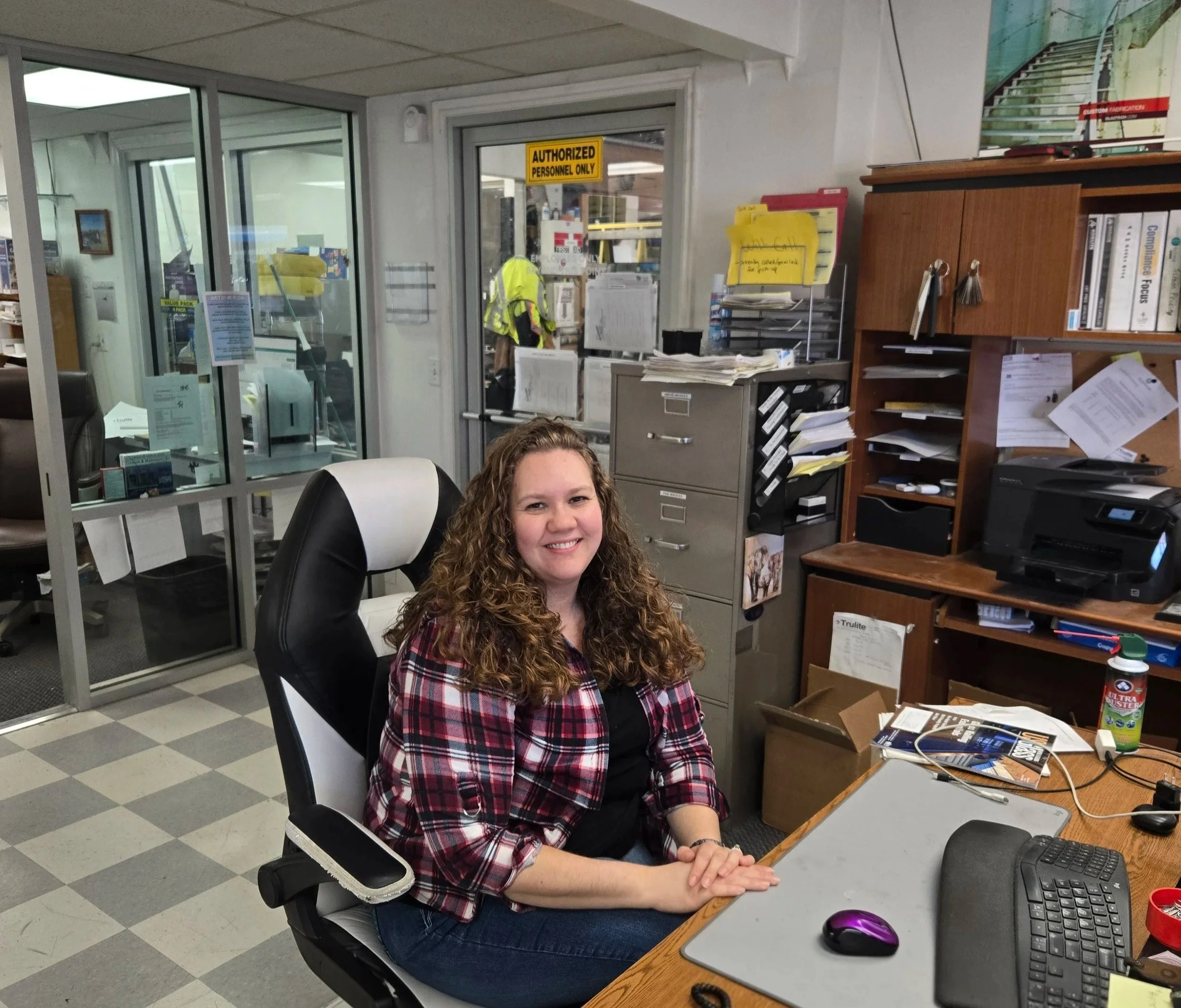 A woman with curly hair smiling and sitting in a black and white office chair at a cluttered desk in an office environment. Behind her are filing cabinets, shelves with papers, and a printer on the desk. Outside the office through the glass door, a person in a yellow safety vest is visible. There is a sign that says 'Authorized Personnel Only' on the door.