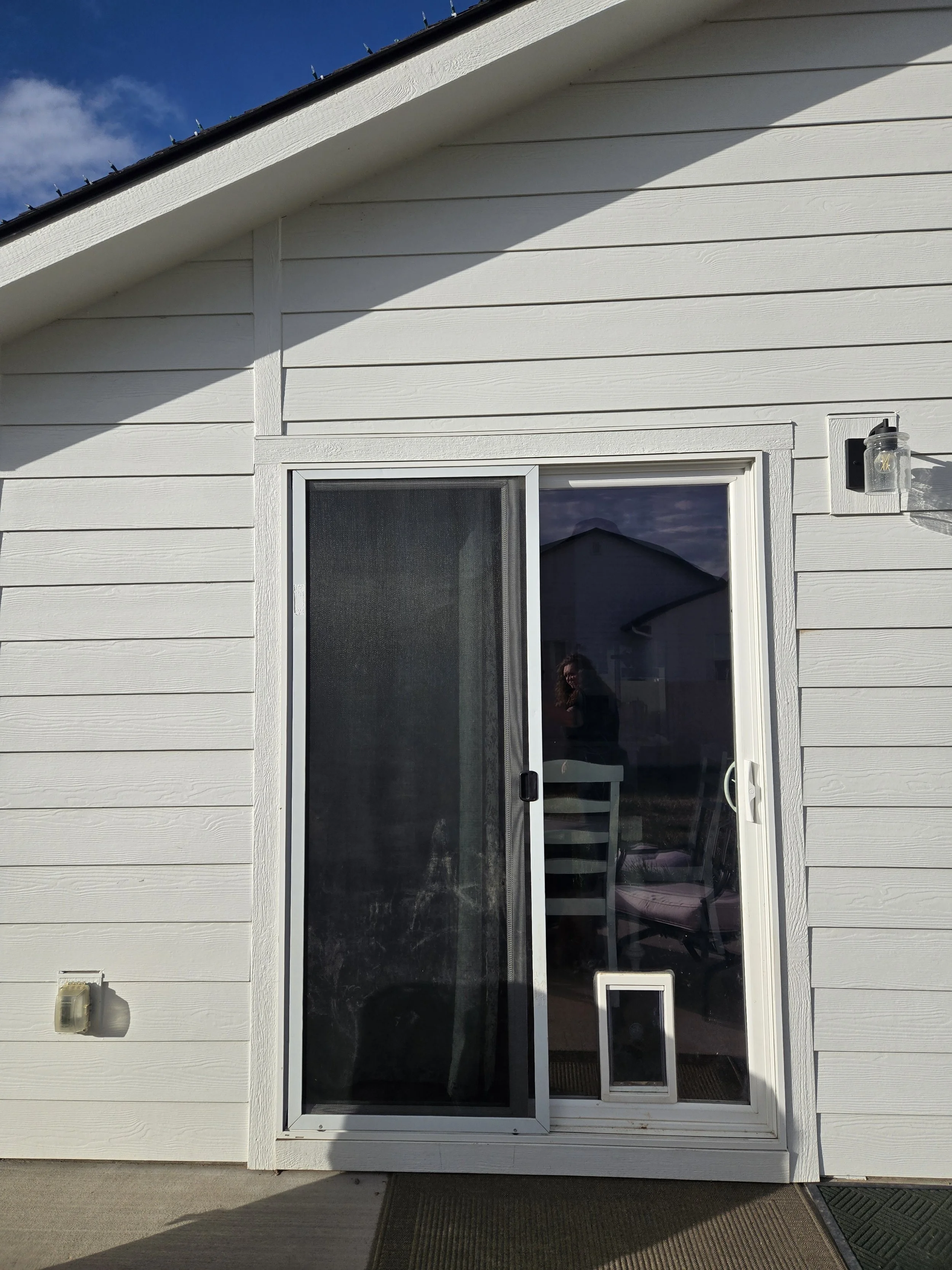 White siding exterior house with a sliding glass door, a screen door partially open, a small pet door at the bottom of the sliding door, and outdoor wall-mounted light fixtures, under a blue sky with some clouds.