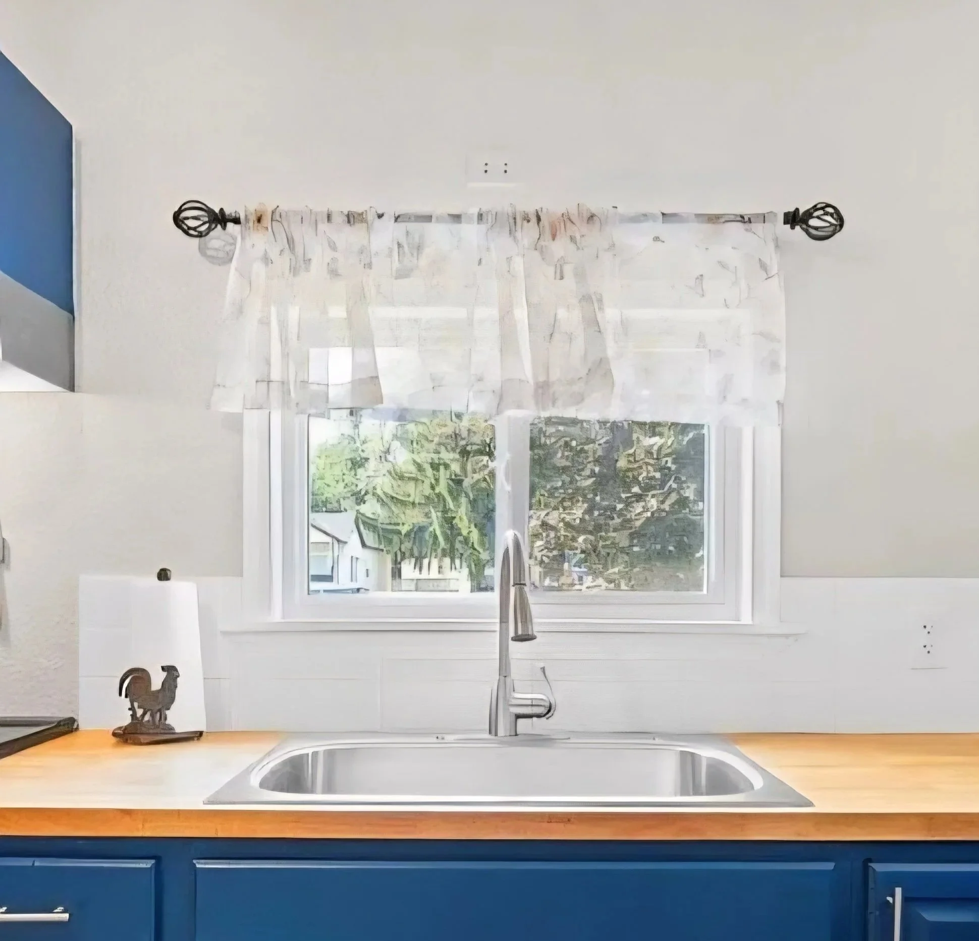 Kitchen sink with stainless steel faucet, wooden countertop, blue cabinets, window with sheer curtains, and a rooster lamp on the left side of the countertop.
