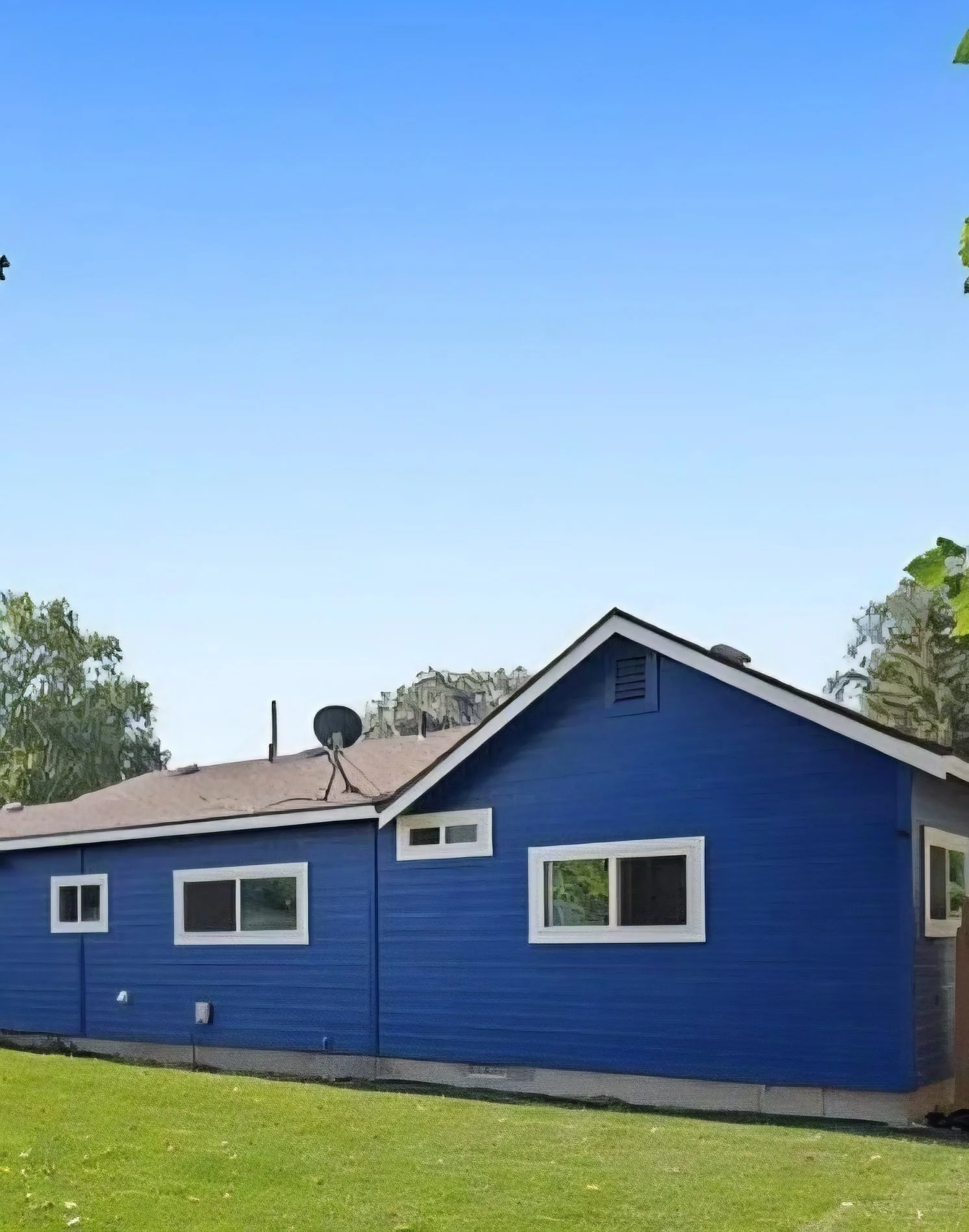 A blue house with white window frames, a sloped roof with satellite dishes, and a green lawn in front, under a clear blue sky.