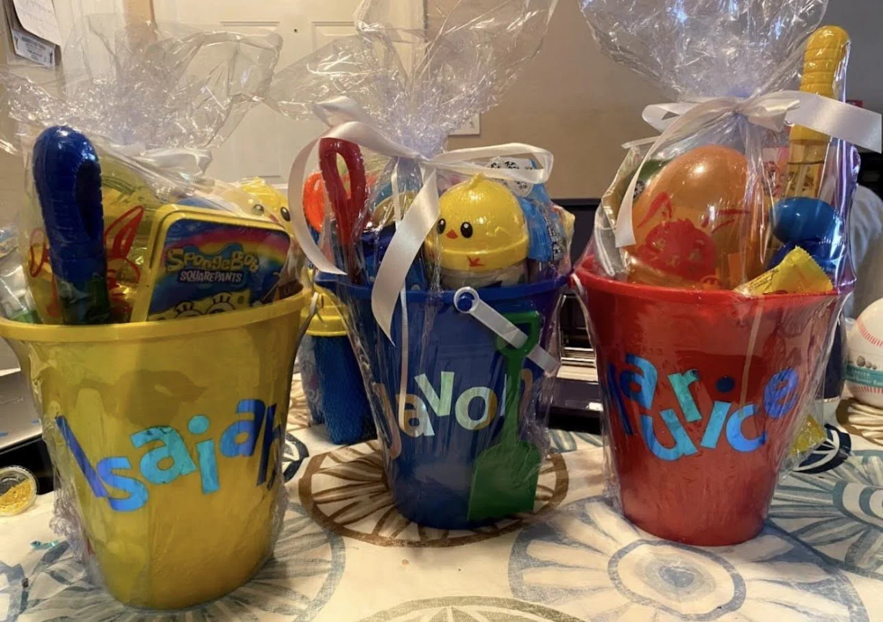 Three colorful buckets wrapped in clear cellophane with bows, filled with toys and goodies, labeled 'fish' in blue letters, set on a table with a patterned tablecloth.