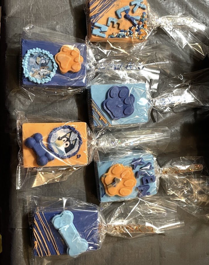 Collection of decorated rice krispy treats wrapped in plastic, featuring bluey, flowers, and alphabet letter toppers in blue and orange colors.