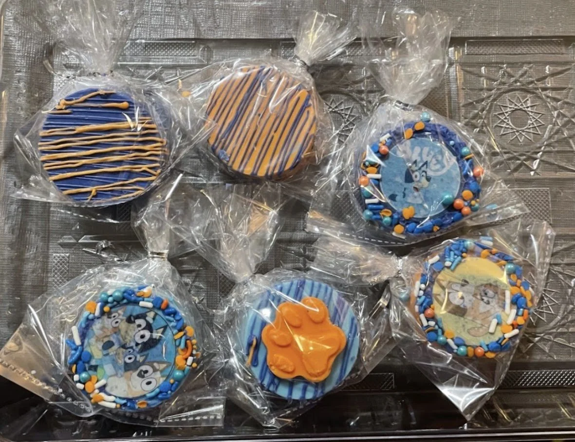 Decorated cookies with blue, orange, and white icing and sprinkles, individually wrapped in plastic bags, placed on a metal tray.