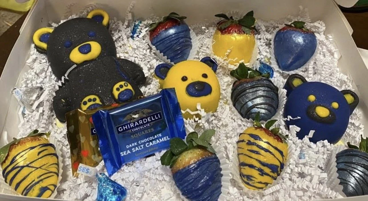 Box of strawberries dipped in blue, yellow, and silver chocolate with bear-shaped and heart-shaped decorations, surrounded by white shredded paper.