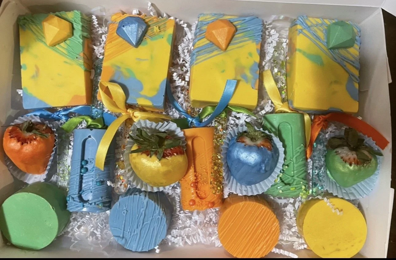 Colorful decorated soap bars shaped like rectangles, circles, and squares with small soap embellishments, arranged in a white box with shredded paper filler.