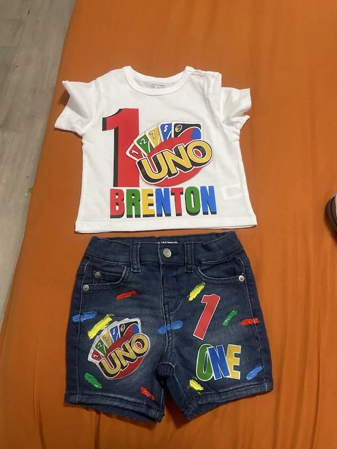 Children's birthday outfit with a white T-shirt and dark denim shorts, featuring colorful UNO cards, the number 1, and the words "Brenton" and "One" in bright colors.