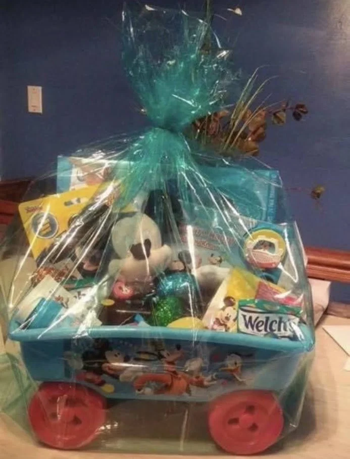 A gift basket wrapped in clear teal plastic, filled with Disney-themed toys and products, with pink wheels, on a wooden surface against a blue wall.
