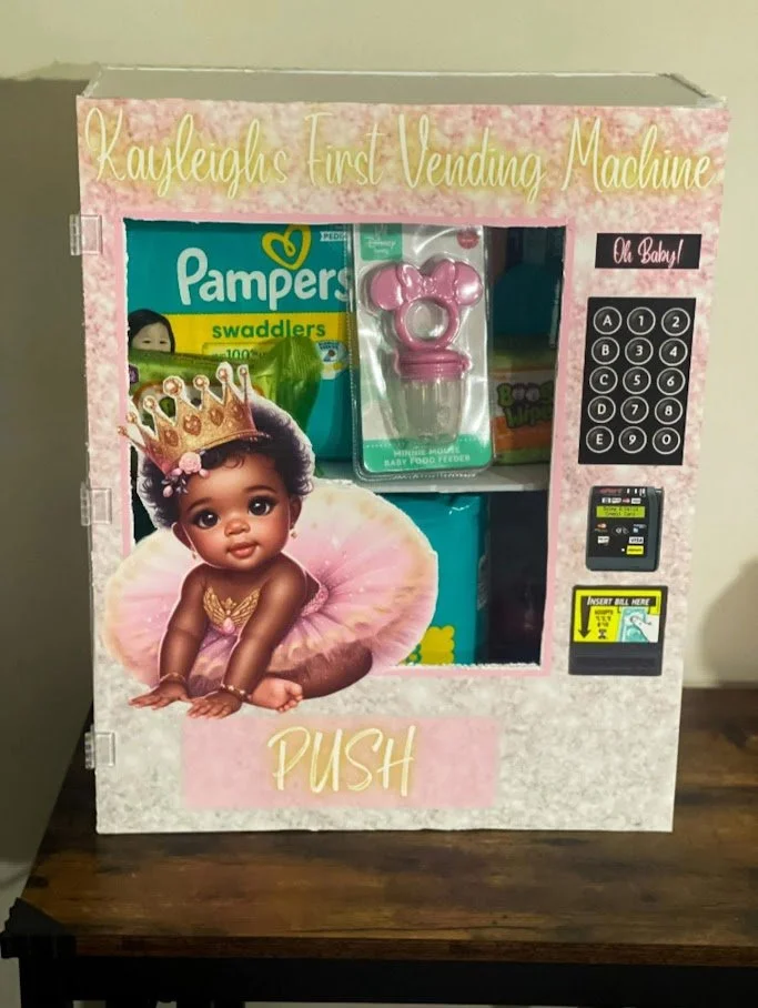 A pink vending machine designed for children called 'Kayleigh's First Vending Machine.' It features a picture of a baby wearing a crown and pink tutu, with the word 'PUSH' on the lower front. The machine includes items like Pampers swaddlers, a pink 