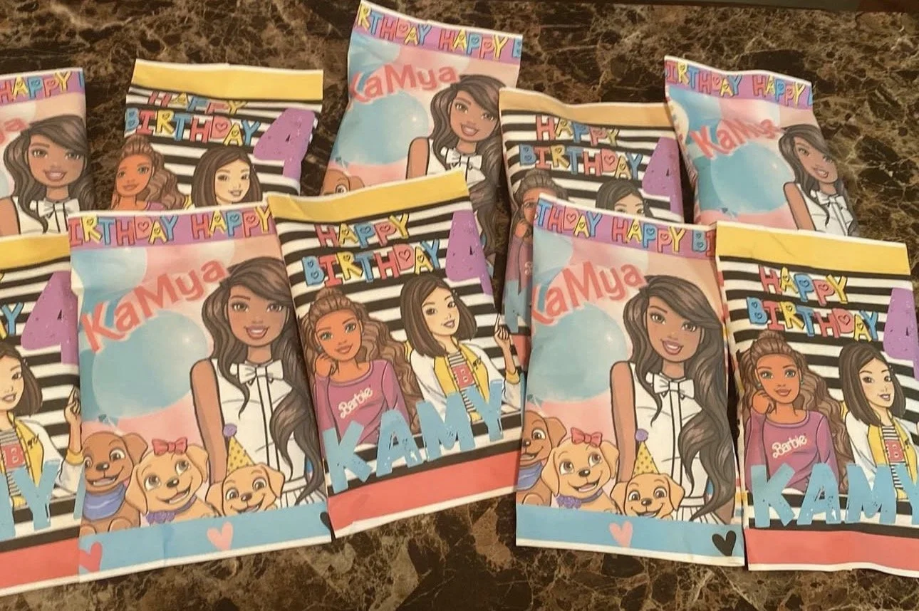 Decorative party favor bags with cartoon girls and puppies, featuring a birthday theme with the name Kamya and the message "Happy Birthday" on a black and white striped background.