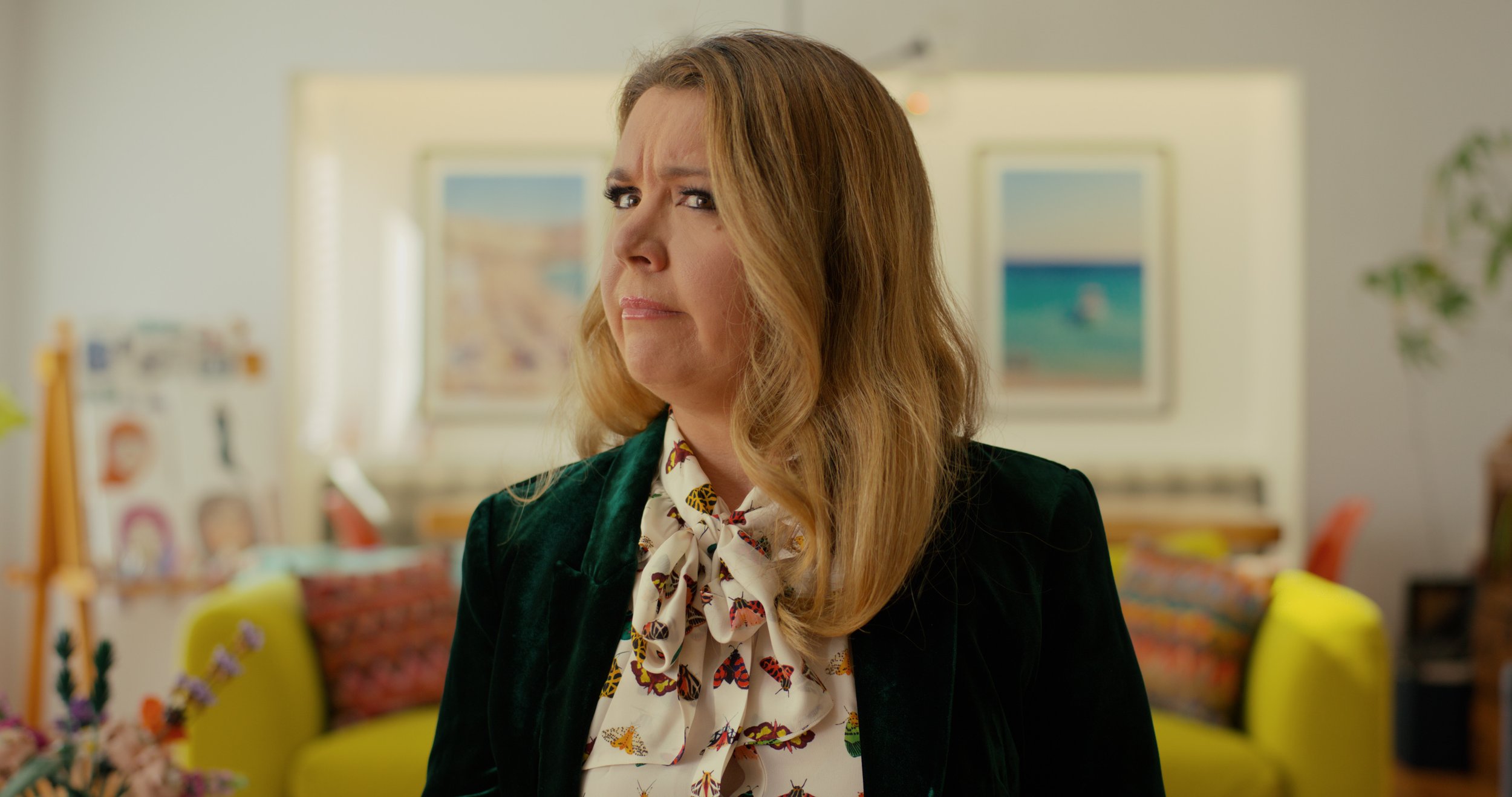 A woman with shoulder-length light brown hair, wearing a dark green jacket and a white blouse with a butterfly pattern, appears puzzled or skeptical inside a colorful, art-filled room.