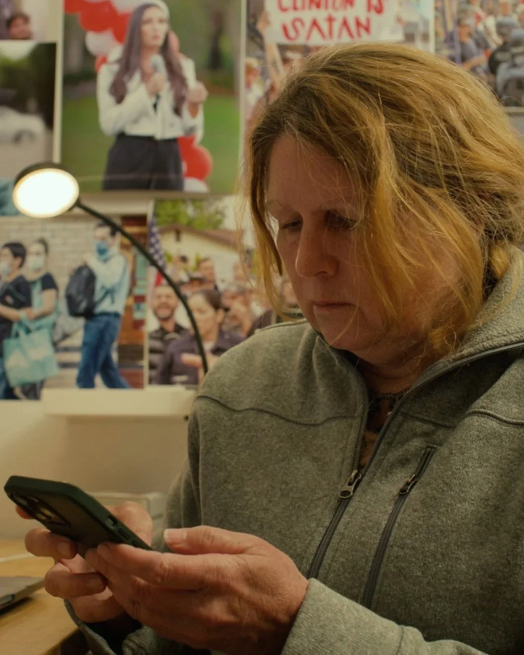 A woman with curly, reddish hair looking at her phone in front of a wall decorated with photos and a sign that reads 'Clinton is Satan.'