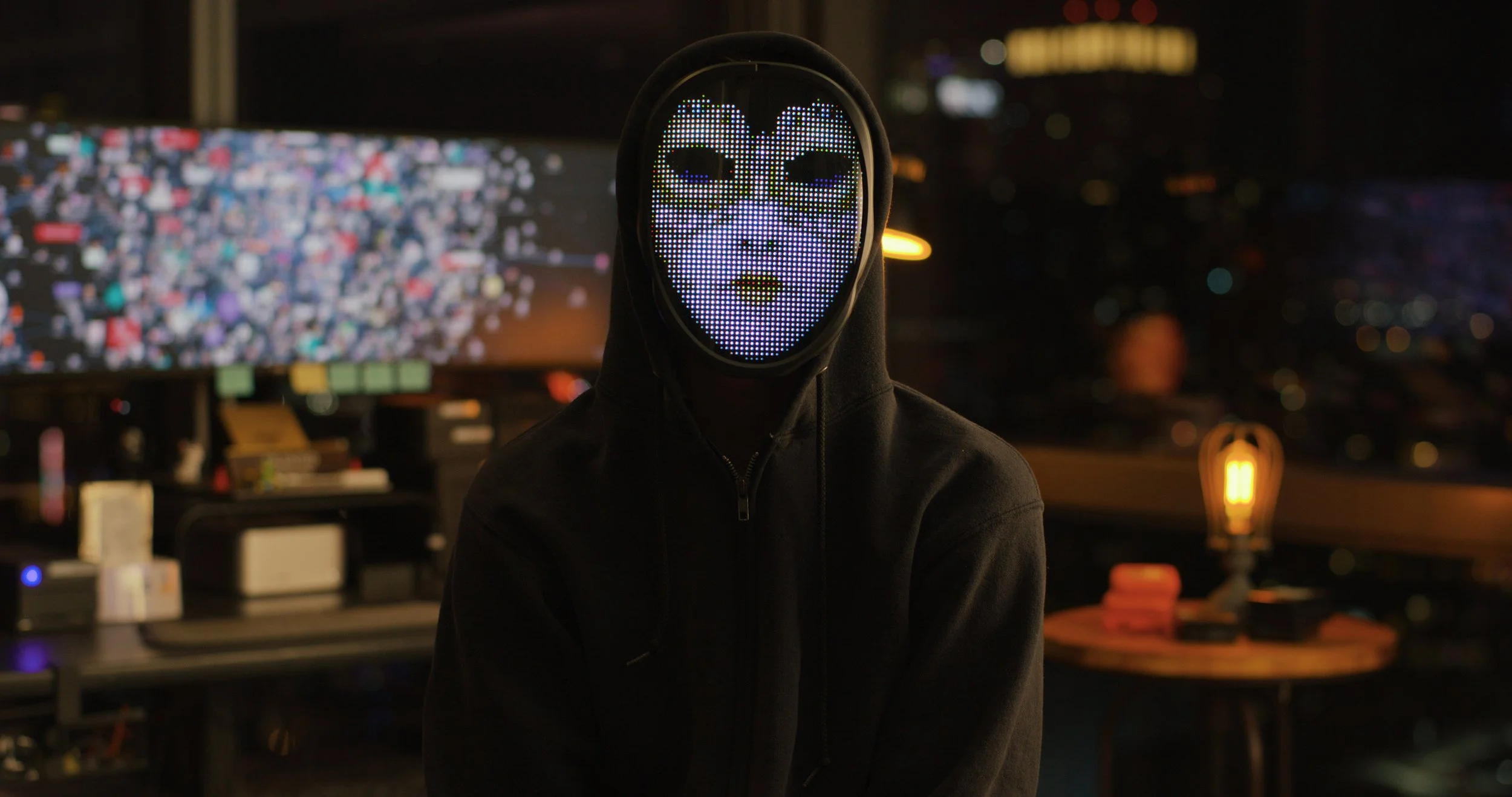 Person wearing a hoodie and a luminous LED mask with a stylized face design, standing indoors at night with city lights and a large screen in the background.