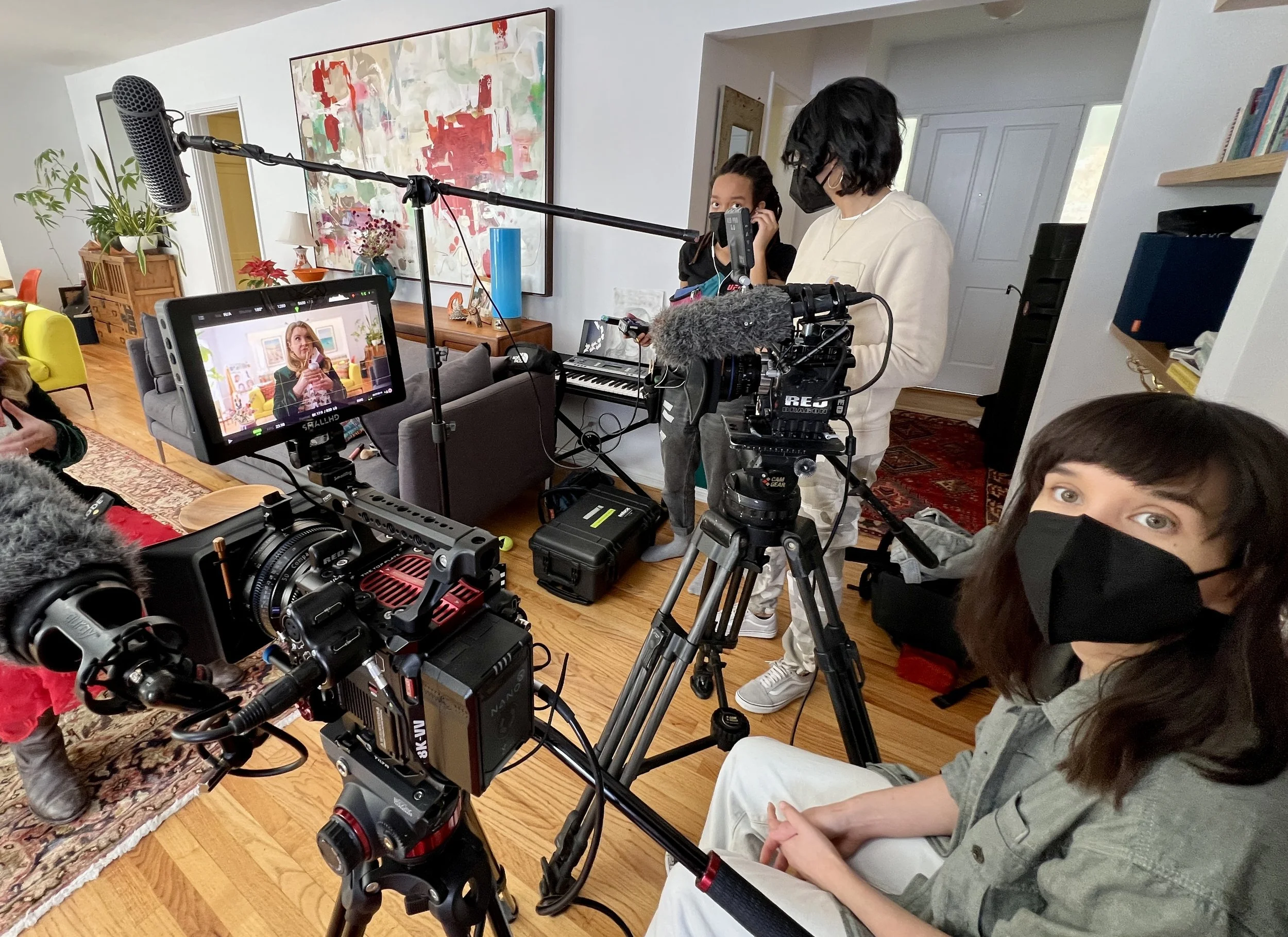 People in a home recording a video or interview, with cameras, microphones, and crew working. A woman is visible on the camera screen, speaking in the background.