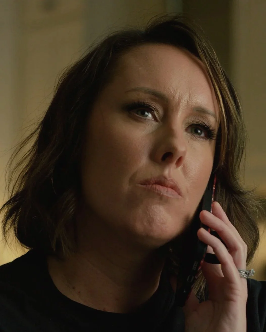 A woman with shoulder-length brown hair, wearing makeup, talking on a cellphone, and wearing a ring on her left hand.