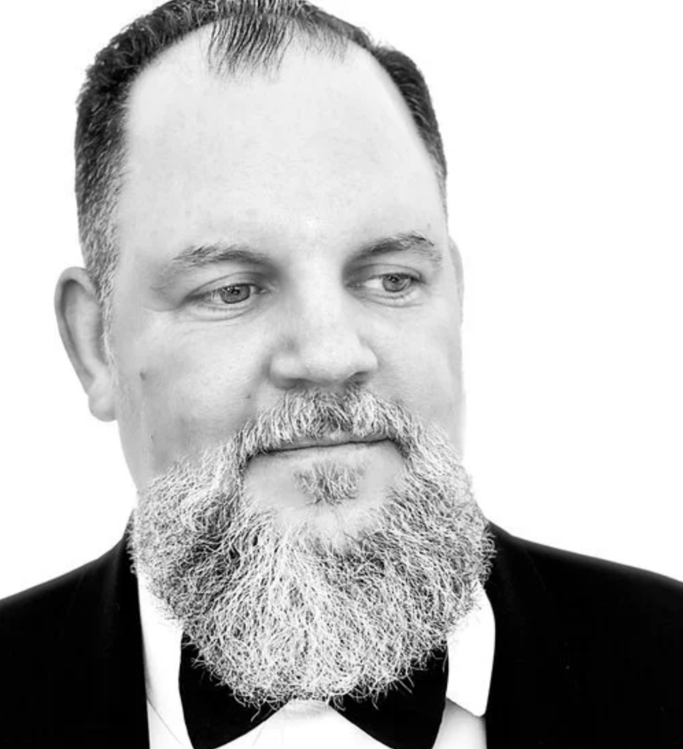 Black and white portrait of a man with a full beard and mustache, wearing a tuxedo with a bow tie, looking slightly to the right.