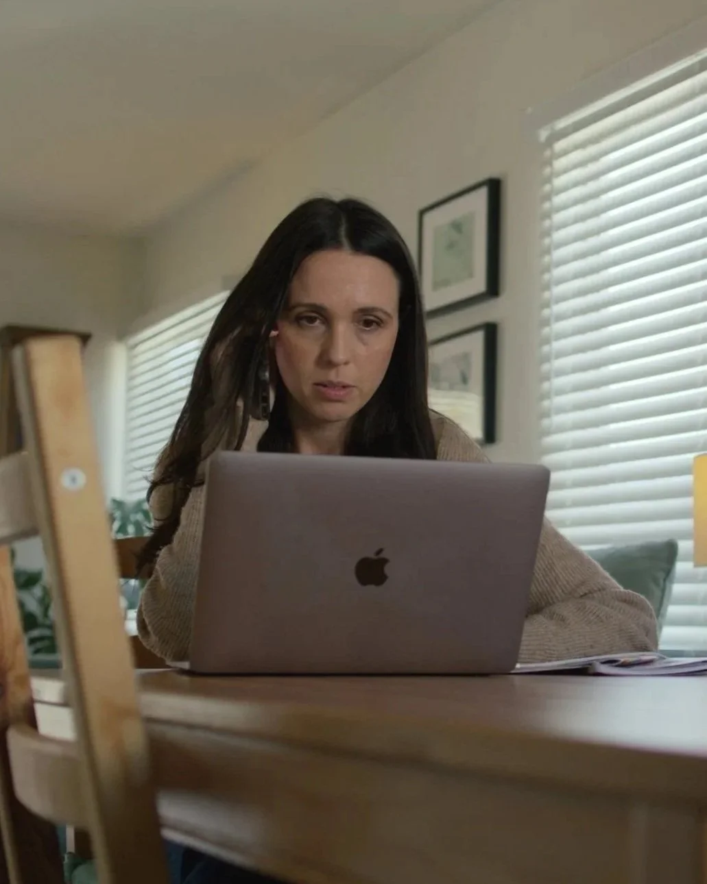A woman with dark hair working on a silver Apple MacBook at a wooden table in a room with framed pictures on the wall and blinds covering the window.