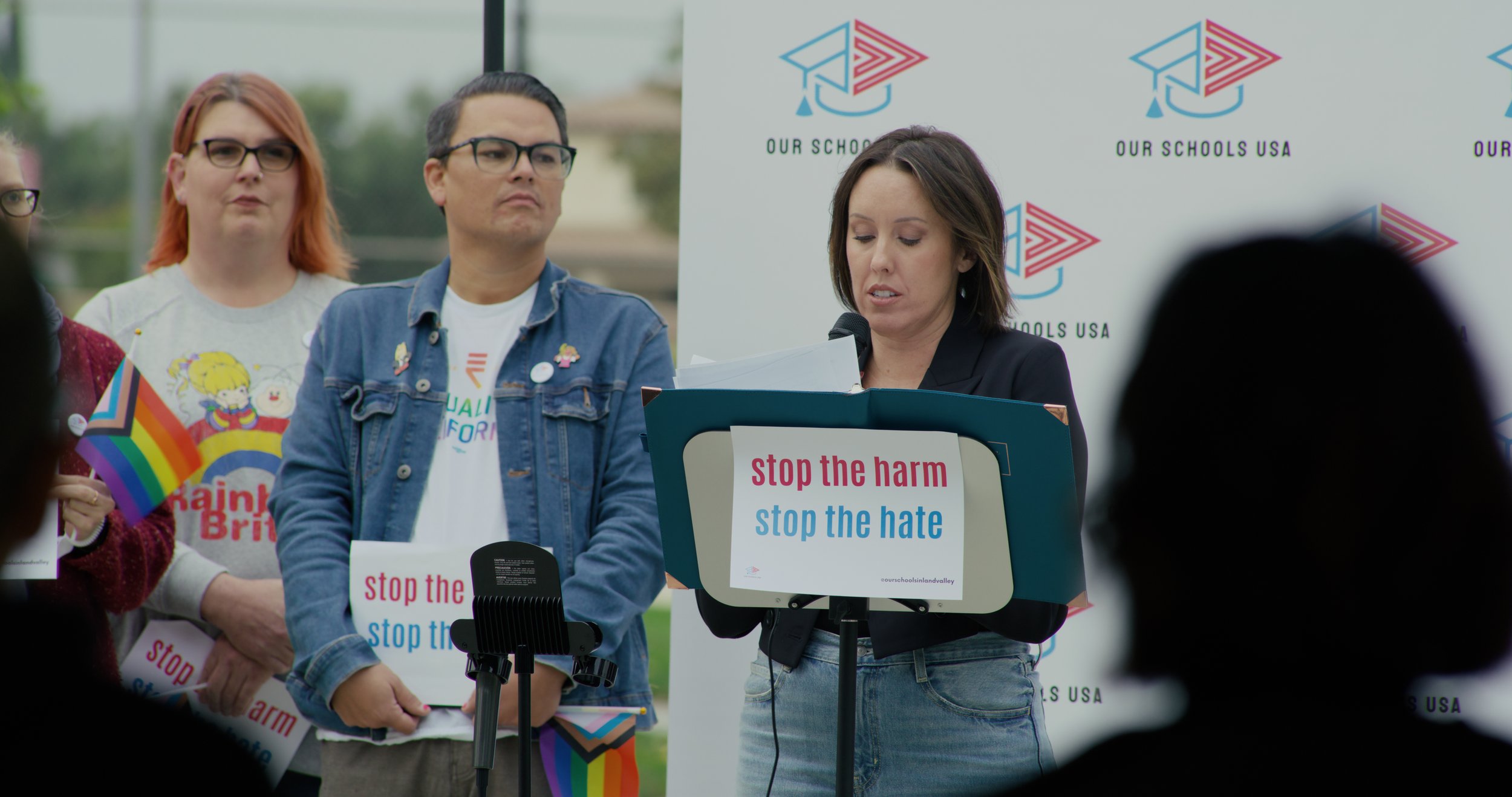 A woman speaking at a podium during a rally or event with a sign that says 'stop the harm, stop the hate.' Several people stand behind her, some holding signs and rainbow flags. There is a backdrop with a logo and the text 'Our Schools USA'.