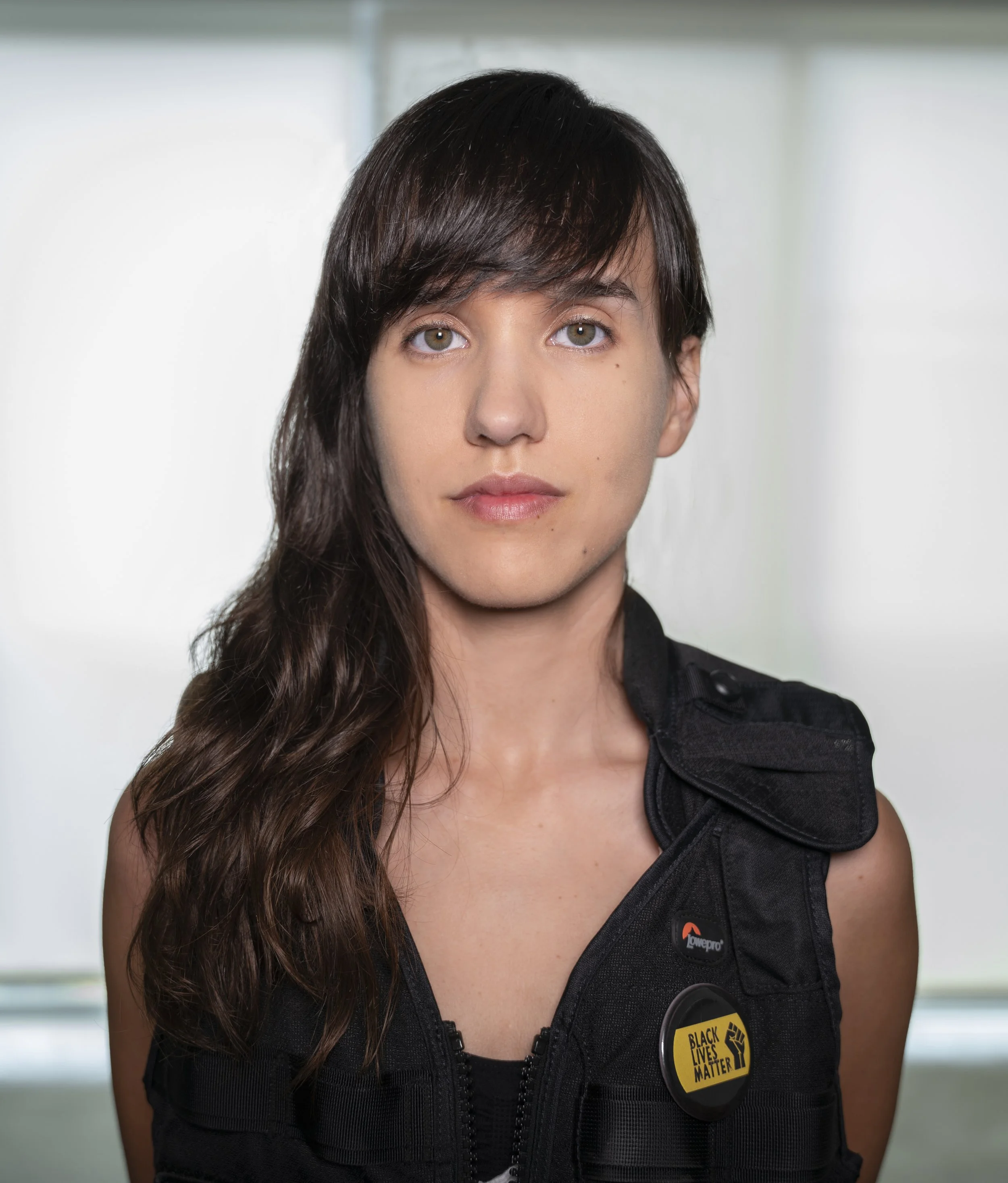 A young woman with long brown hair and light skin looking directly at the camera, wearing a black vest with a 'Black Lives Matter' button and a 'Lowepro' logo, standing in front of a blurred background with natural lighting.