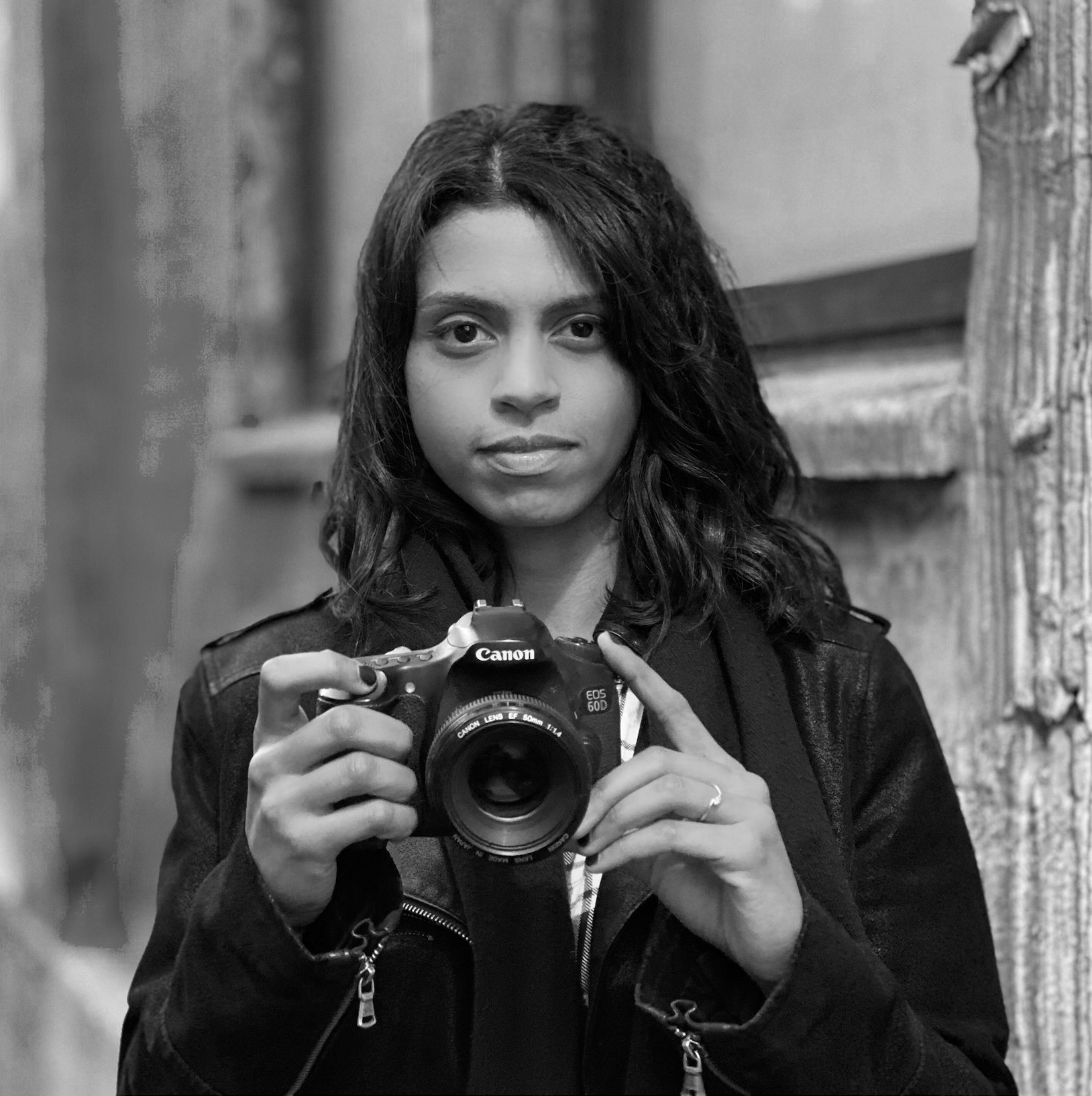 A woman with dark hair holding a Canon EOS 60D camera, looking at the mirror, standing in front of a textured wooden wall.