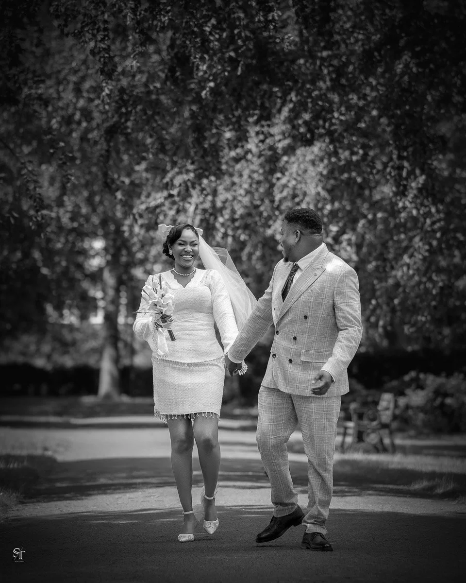 A black and white photograph of a smiling bride and groom holding hands and walking in a park. The bride is wearing a white dress, veil, and heels, while the groom is dressed in a light-colored suit and dark shoes.