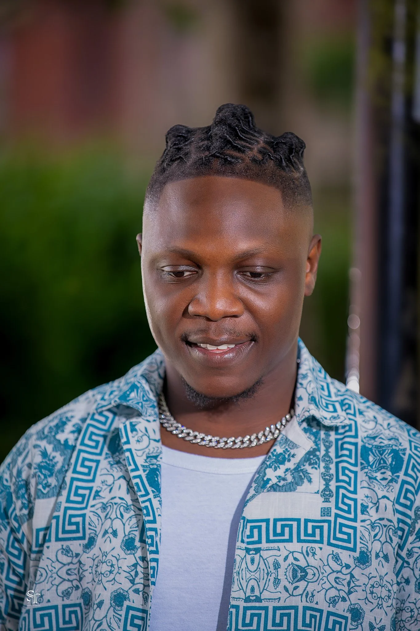 A young man with braided hairstyle, wearing a patterned blue and white shirt, a white T-shirt, and a silver chain necklace, looking down with a slight smile.