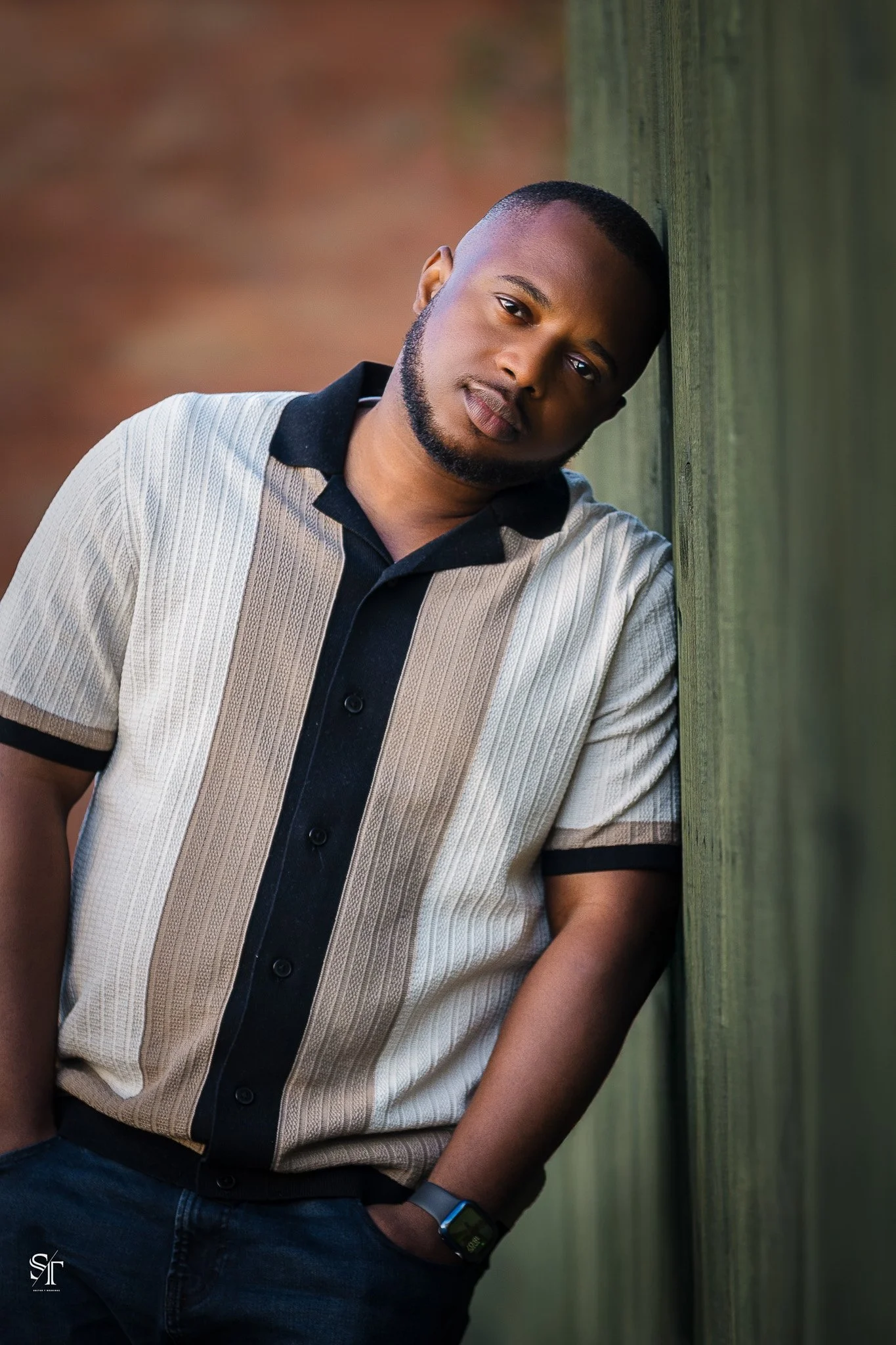 A young man with a beard, wearing a beige and black striped short-sleeve shirt, standing with his back against a green wooden wall, looking at the camera.