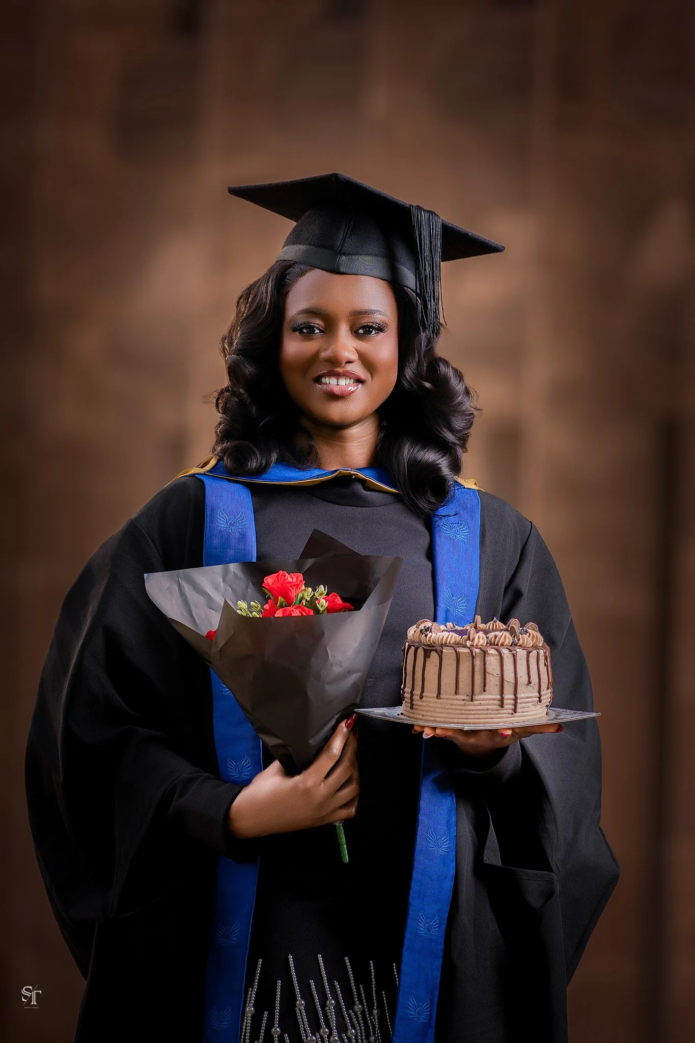 A woman in graduation attire holding a bouquet of flowers and a chocolate cake with chocolate drizzle.