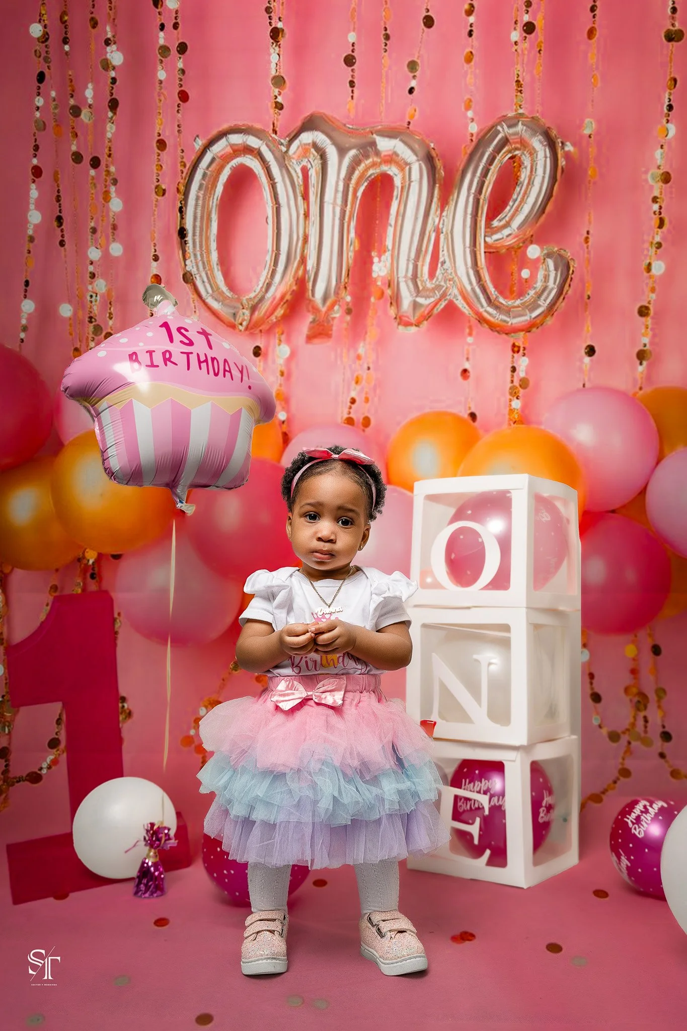 A young girl celebrating her first birthday in a pink-themed party with balloons, a cupcake-shaped balloon with '1st Birthday!' written on it, and a backdrop with 'one' in balloon letters.