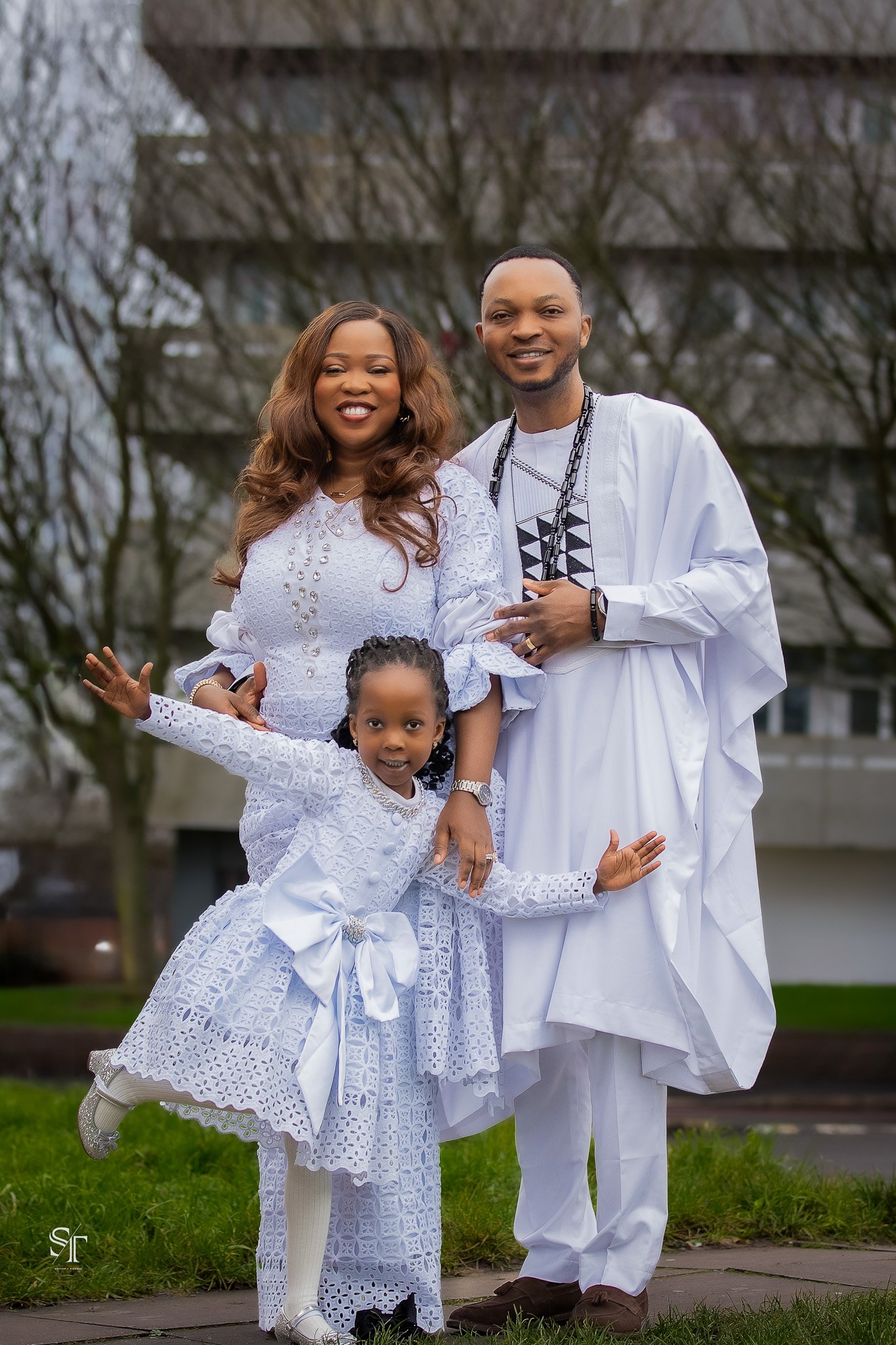 A family of four dressed in matching white attire outdoors with trees and a building in the background.