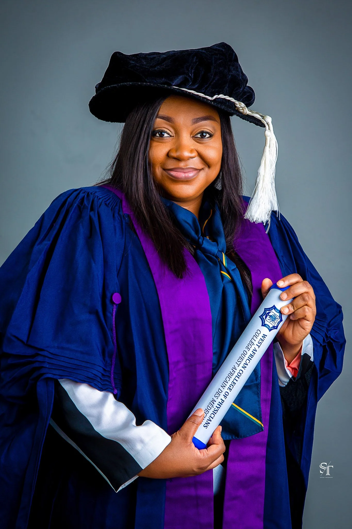 A woman in graduation attire, wearing a black cap with a white tassel and a blue gown with purple accents, holding a diploma in her hand and smiling.